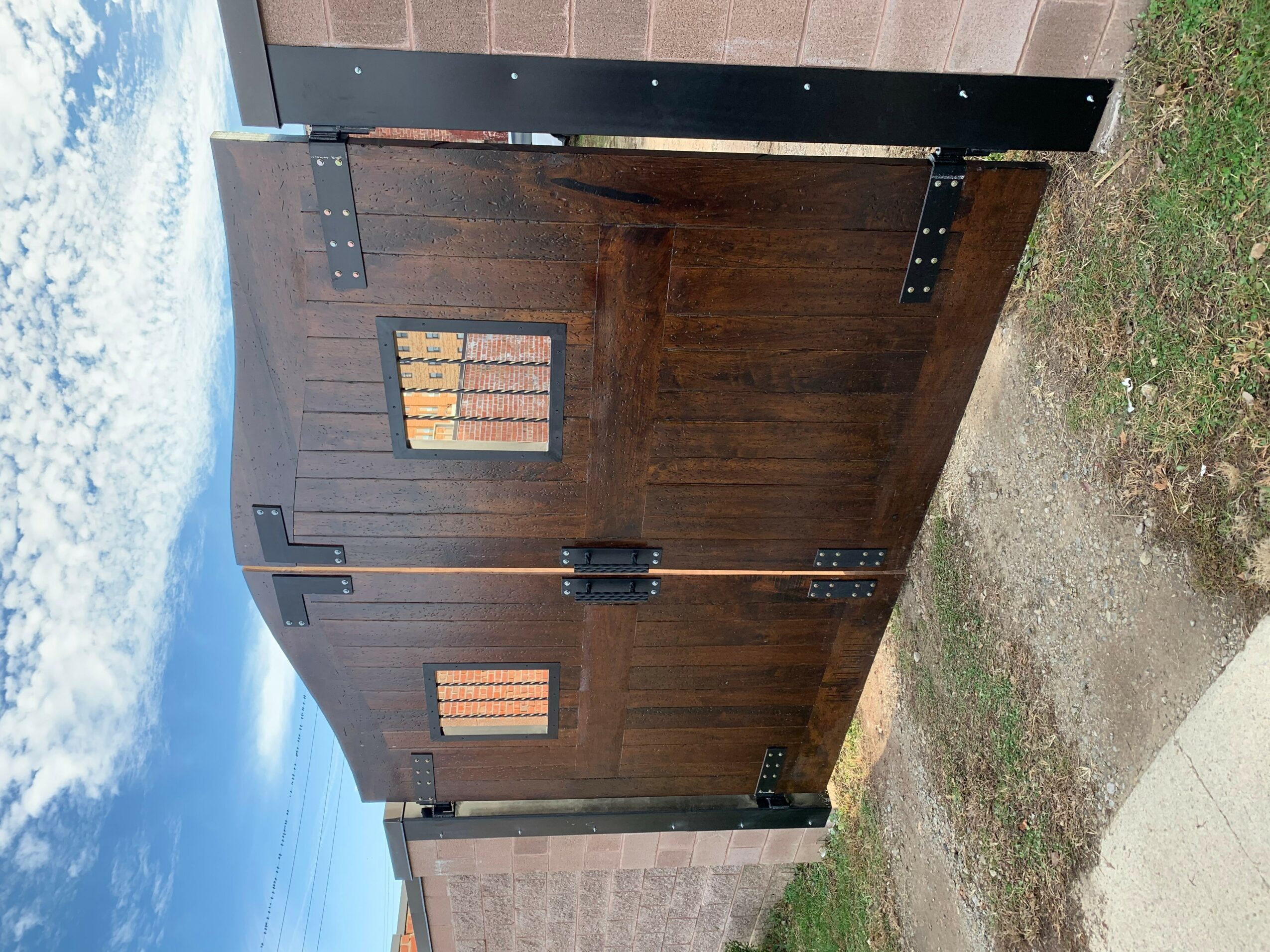 Wooden gate with black hinges and latch, two small windows, set against a cloudy sky and grassy ground.