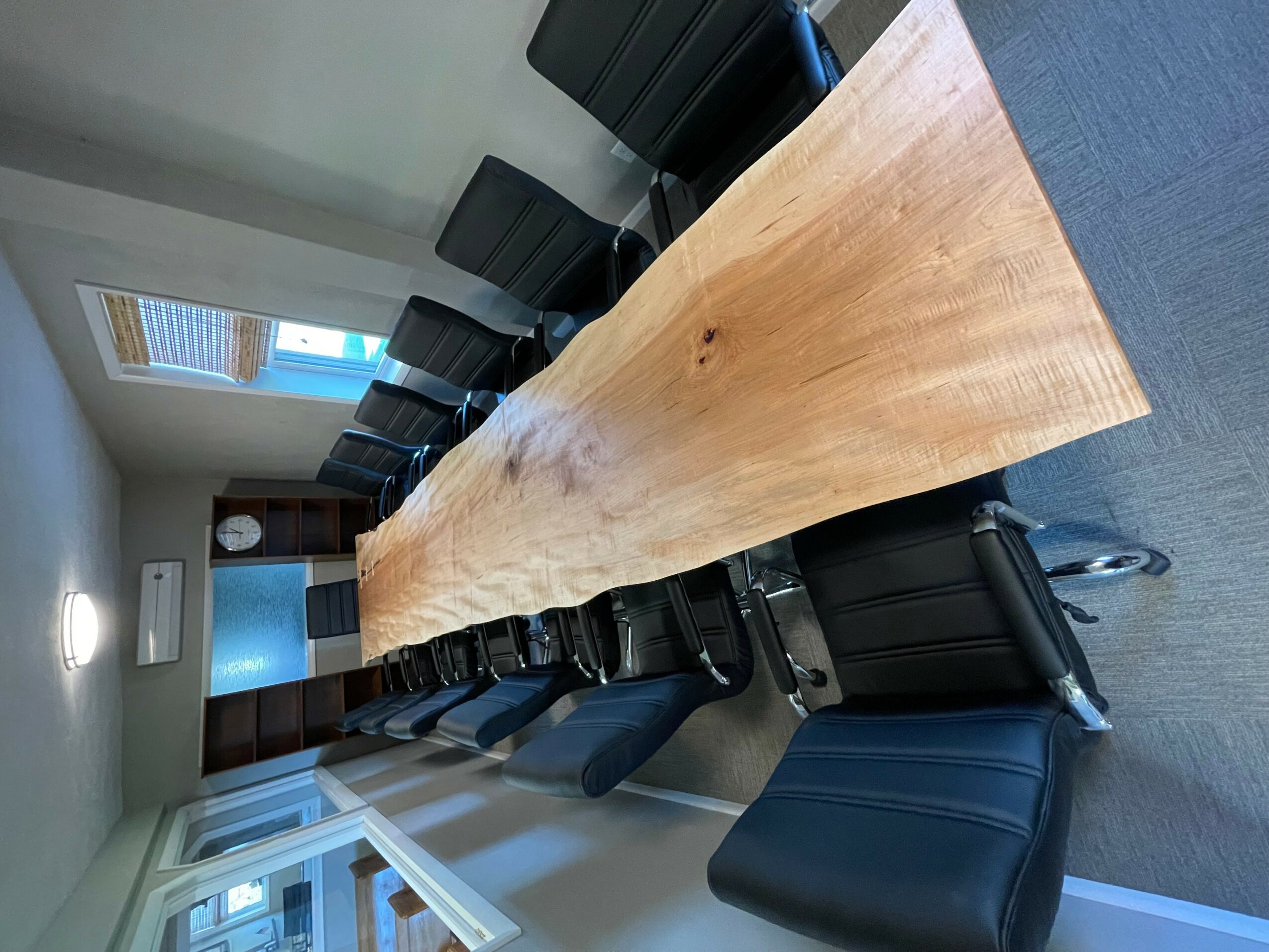 Long wooden table with black chairs in a room with windows and a door, viewed from the side.