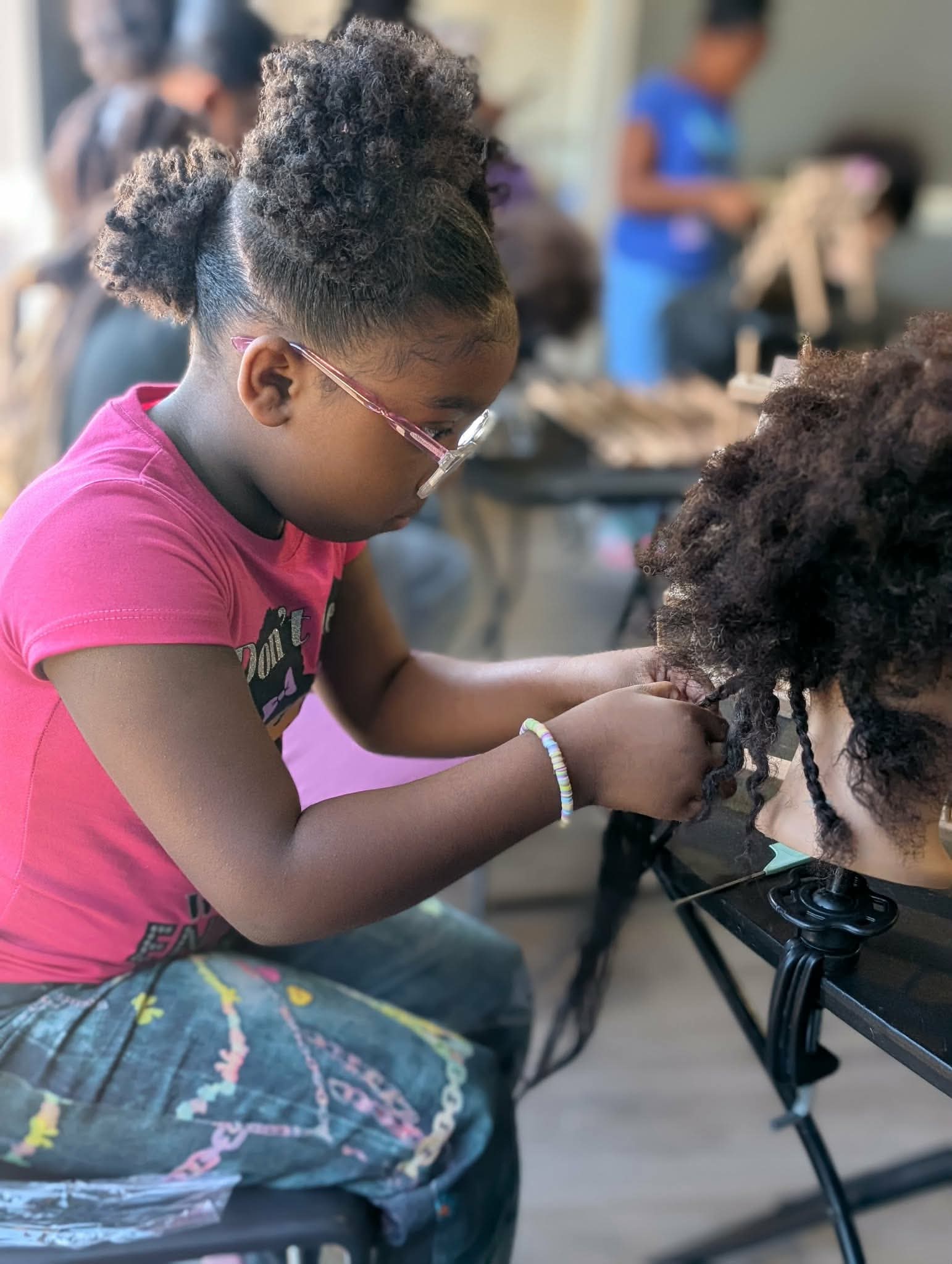 Girl with glasses and curly hair braiding another person's hair in a classroom or salon setting.