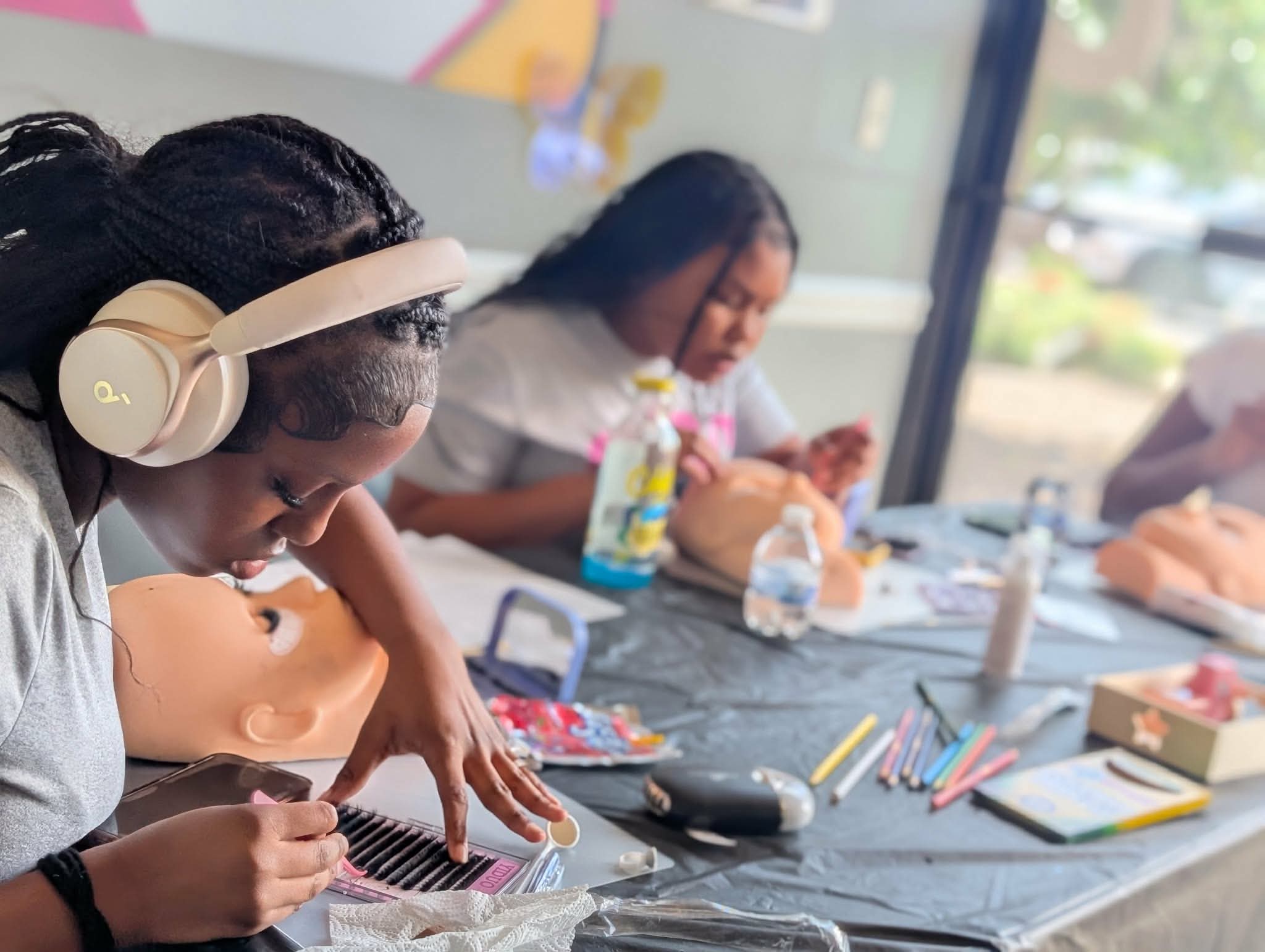 Two girls sit at a table with art supplies, one wearing headphones and focusing on a project, in a bright room.