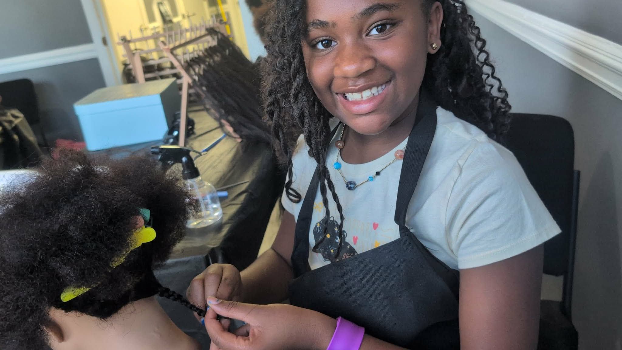 Young girl smiling, wearing a white shirt and apron, sitting at a table with hair styling tools, in a salon.