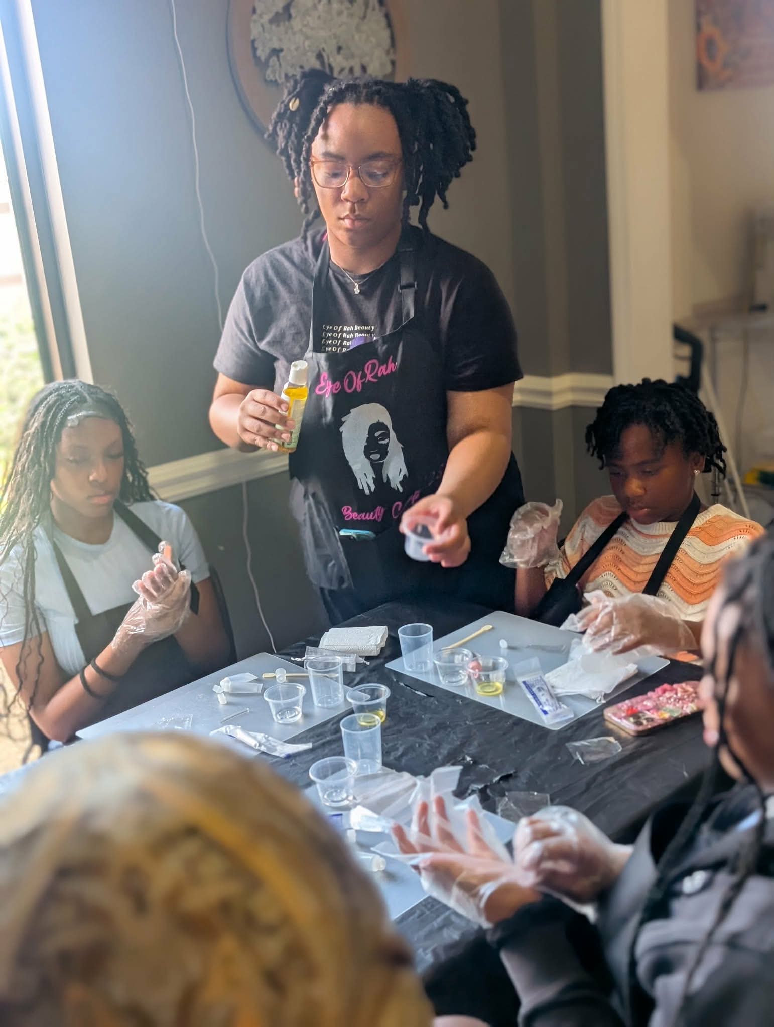 Woman demonstrating a craft to two children at a table with supplies, in a room with a window and light-colored walls.