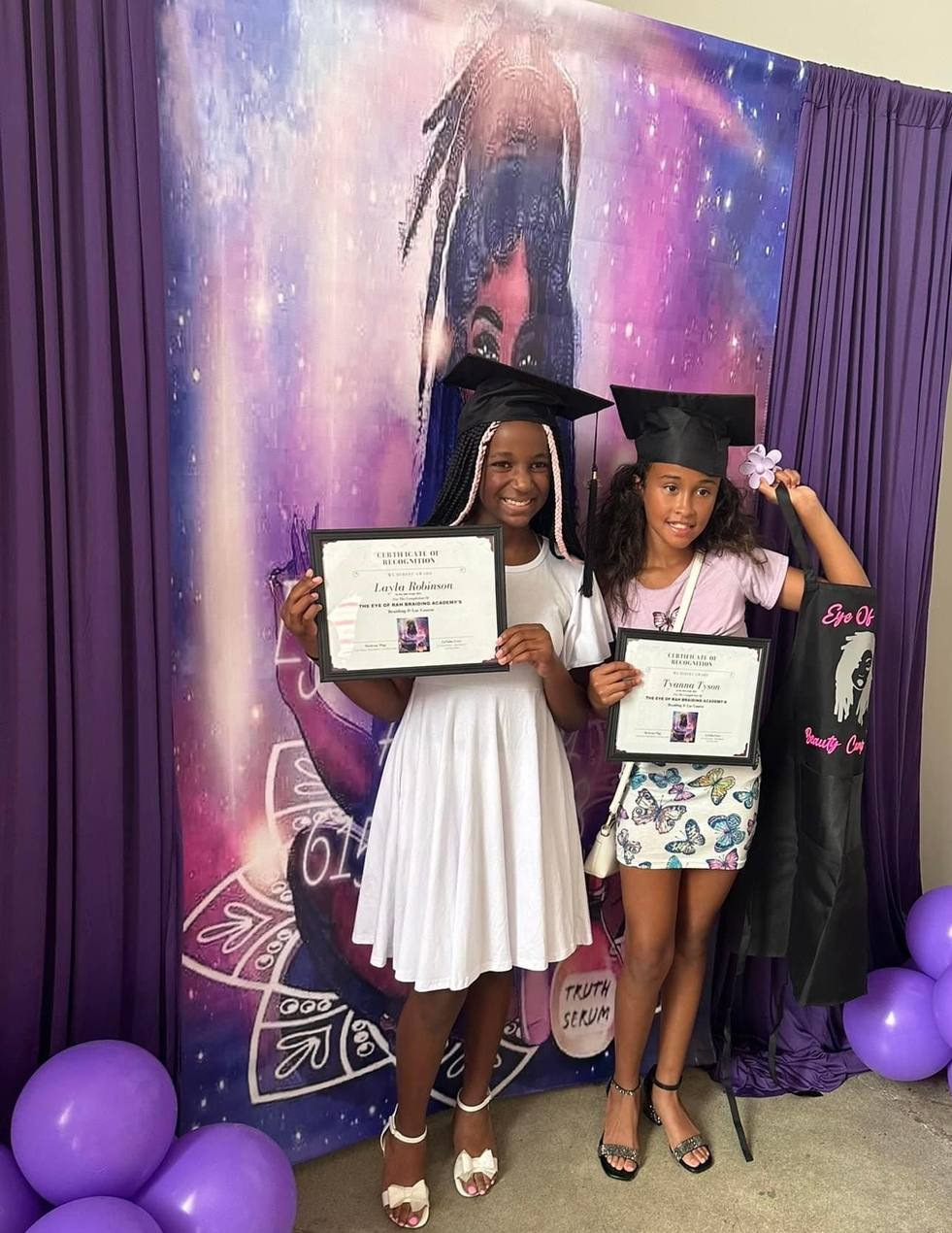 Two young girls in graduation caps and gowns holding diplomas, standing in front of a colorful backdrop with purple balloons nearby.