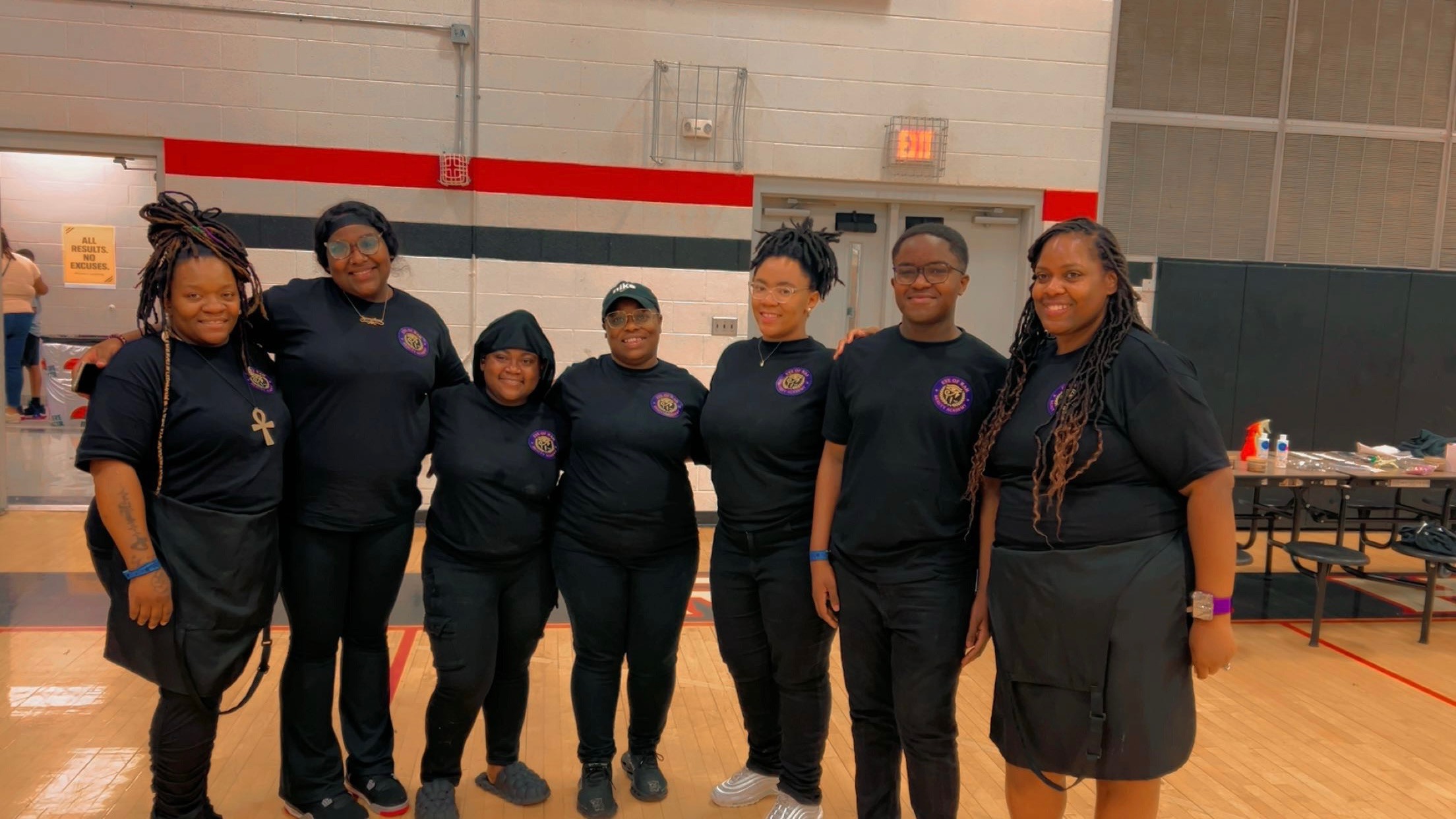Group of seven people standing together indoors, wearing matching dark shirts, smiling, with tables and equipment in the background.