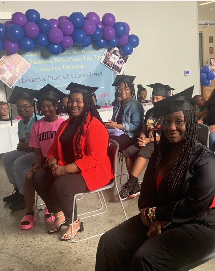Group of graduates in caps and gowns seated indoors with purple balloon decorations behind them.
