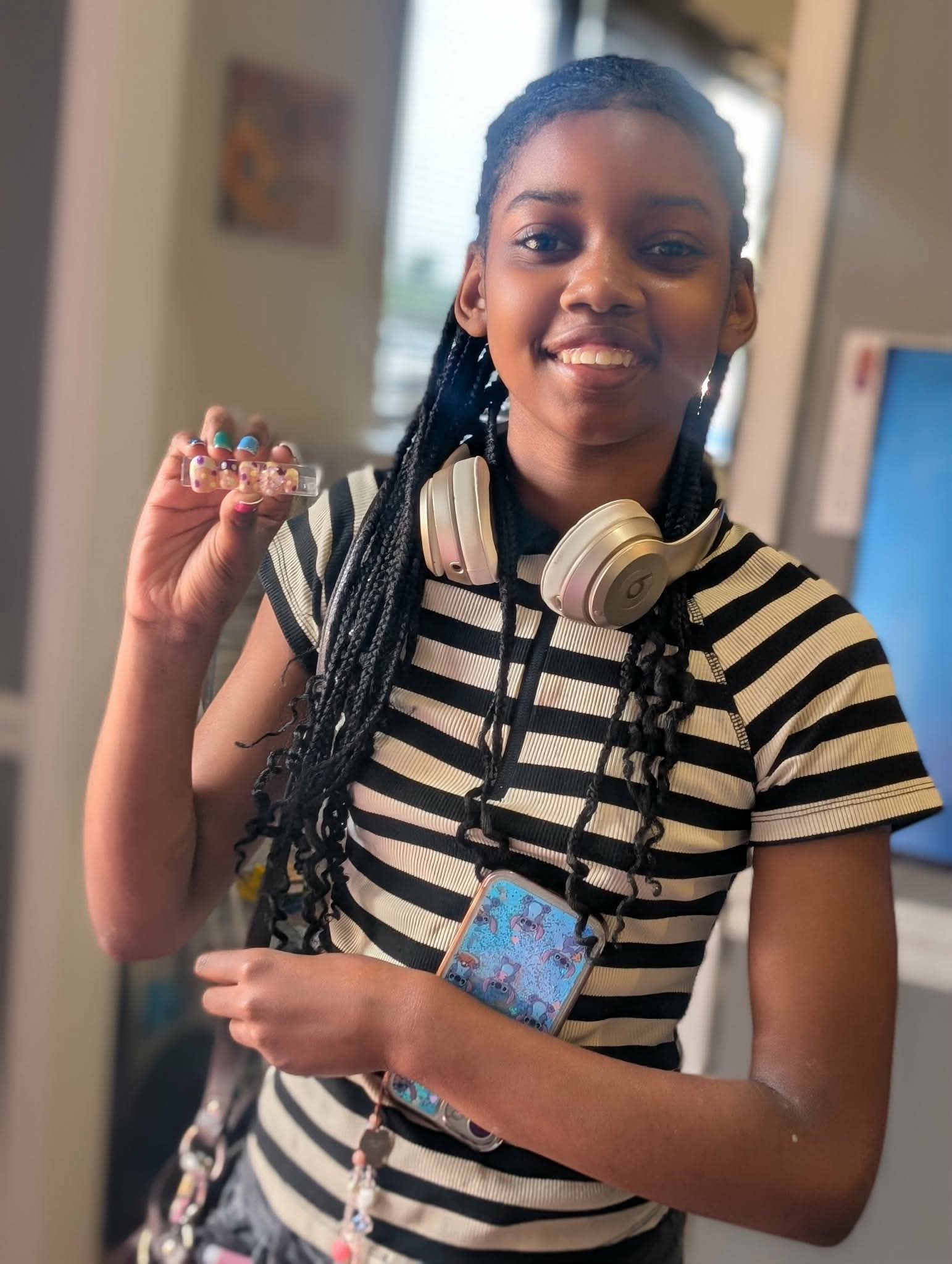 Smiling girl with braided hair holding a small object, wearing headphones around neck and striped shirt, indoors.
