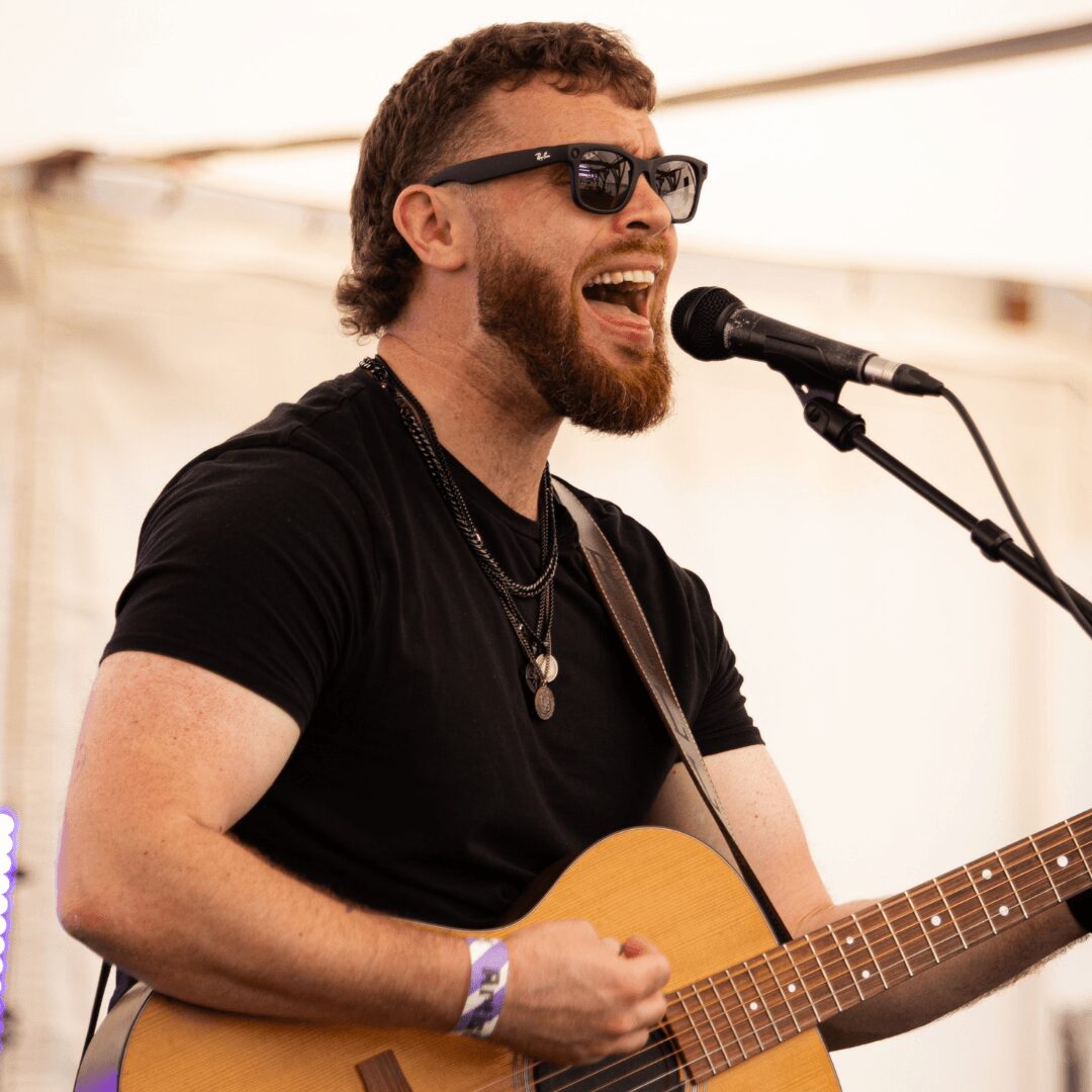 Man with sunglasses singing into a microphone while playing an acoustic guitar, wearing a black shirt and jewelry.