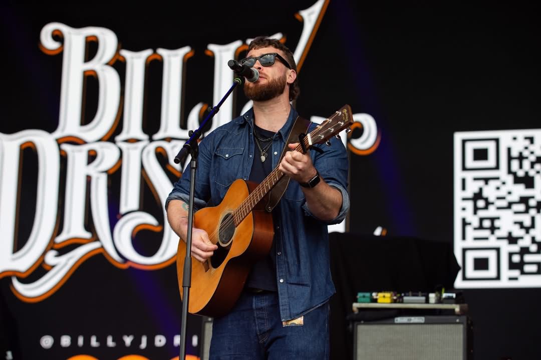 Male musician with sunglasses playing guitar on stage, with a large sign and QR code in background.