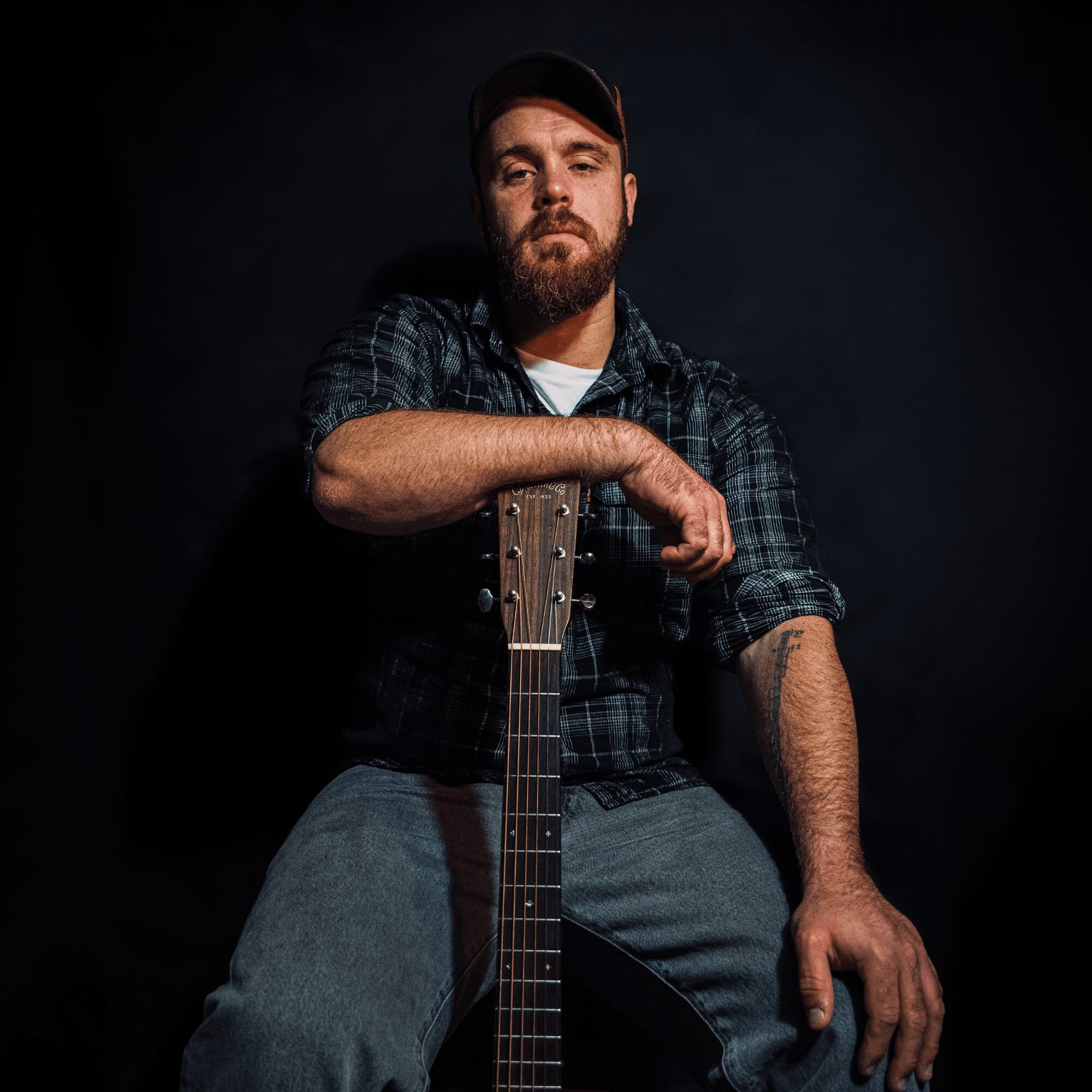 Man with beard and cap holding guitar, sitting against dark background.