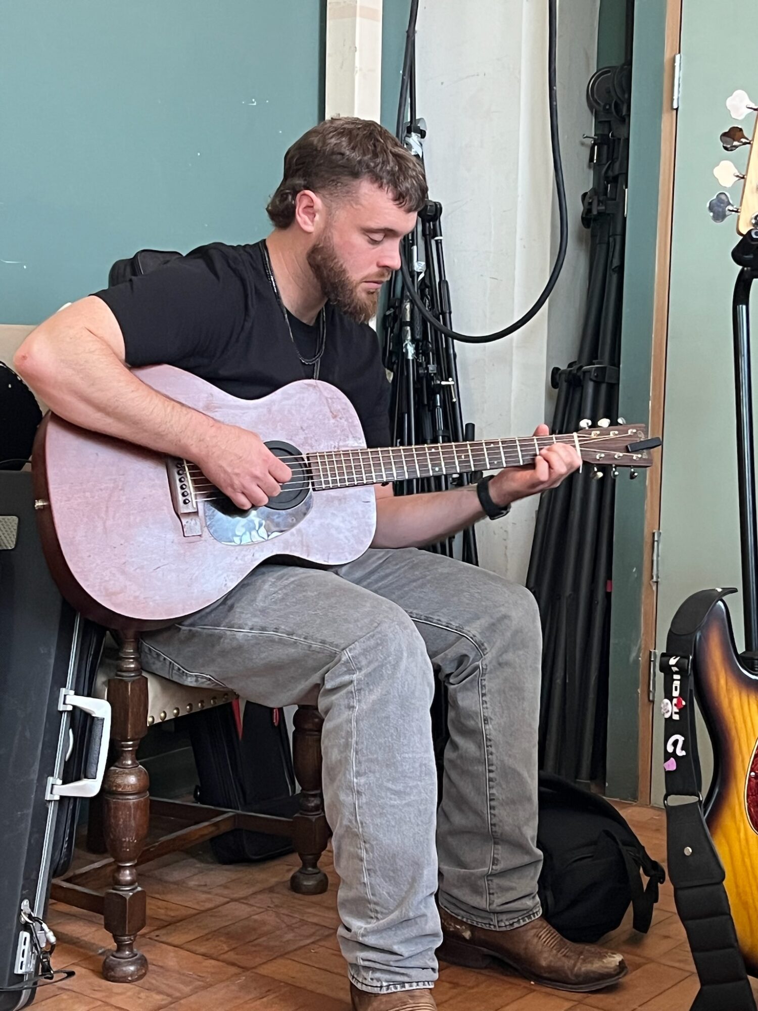 Man with beard playing acoustic guitar while sitting on a wooden chair in a room with musical equipment.