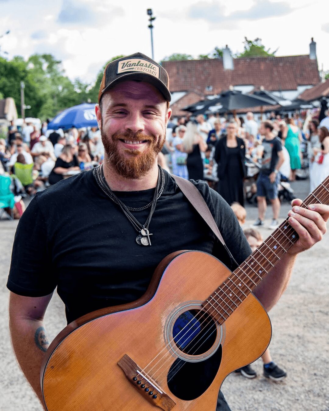 Man with a beard and cap smiling, holding an acoustic guitar at an outdoor event with a crowd and buildings in background.