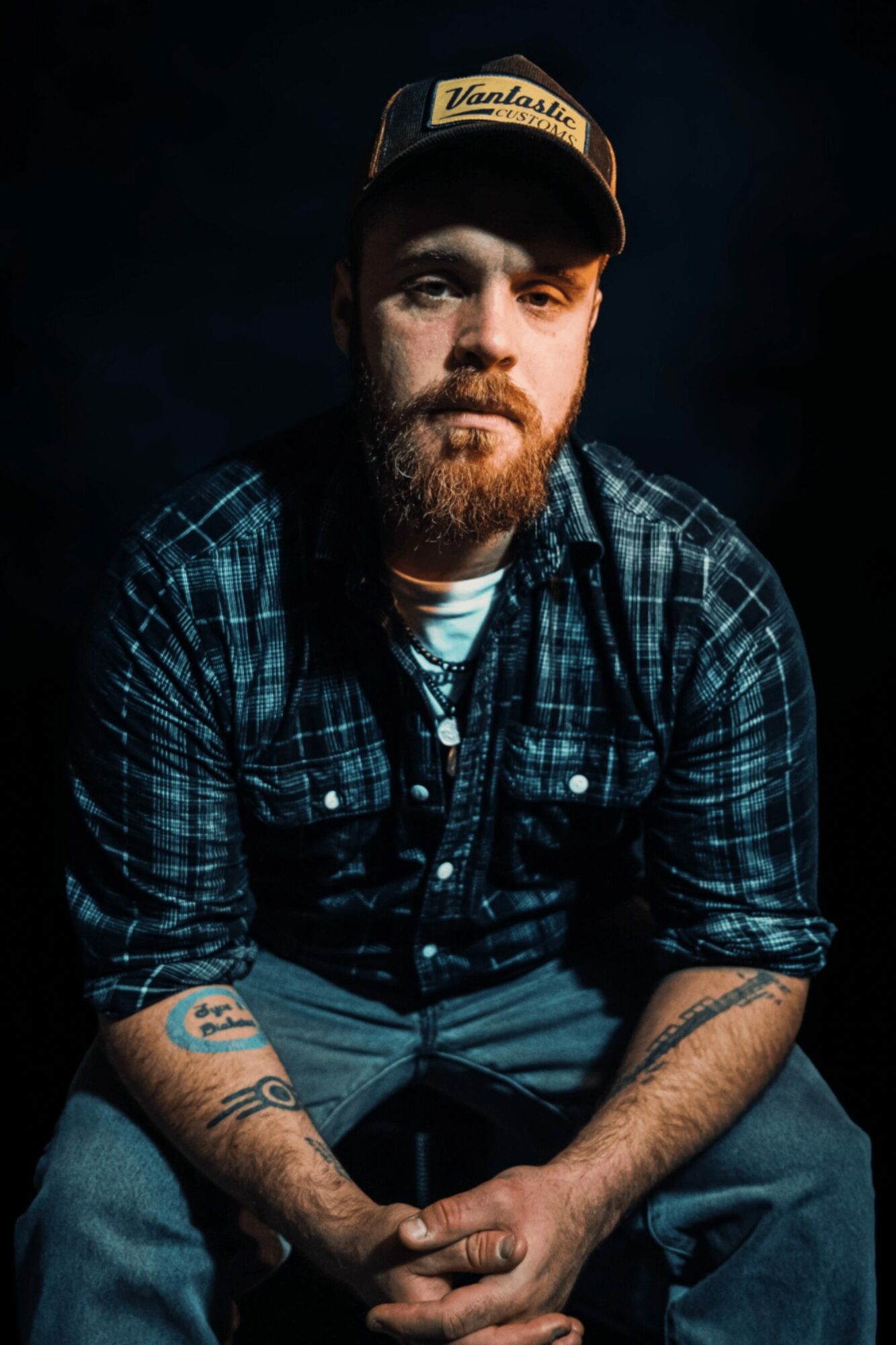 Man with a beard wearing a cap and plaid shirt, sitting with hands clasped, dark background.