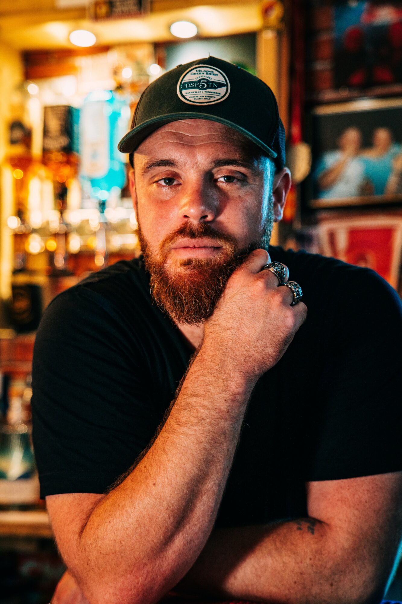 Man with a beard and black cap, resting his chin on his hand in a dimly lit bar or restaurant.