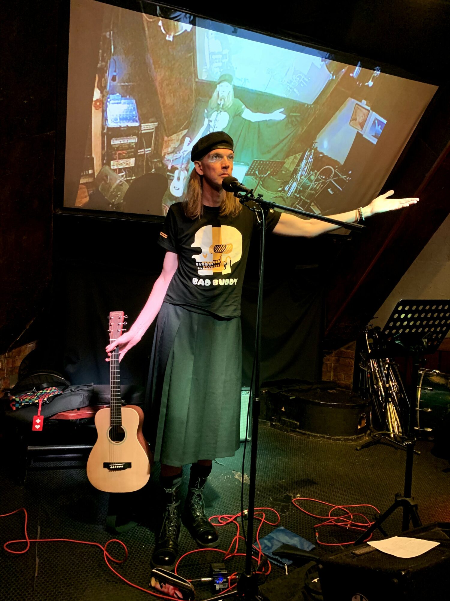 Woman performing on stage with microphone, guitar, and a large screen behind her, in a music venue.