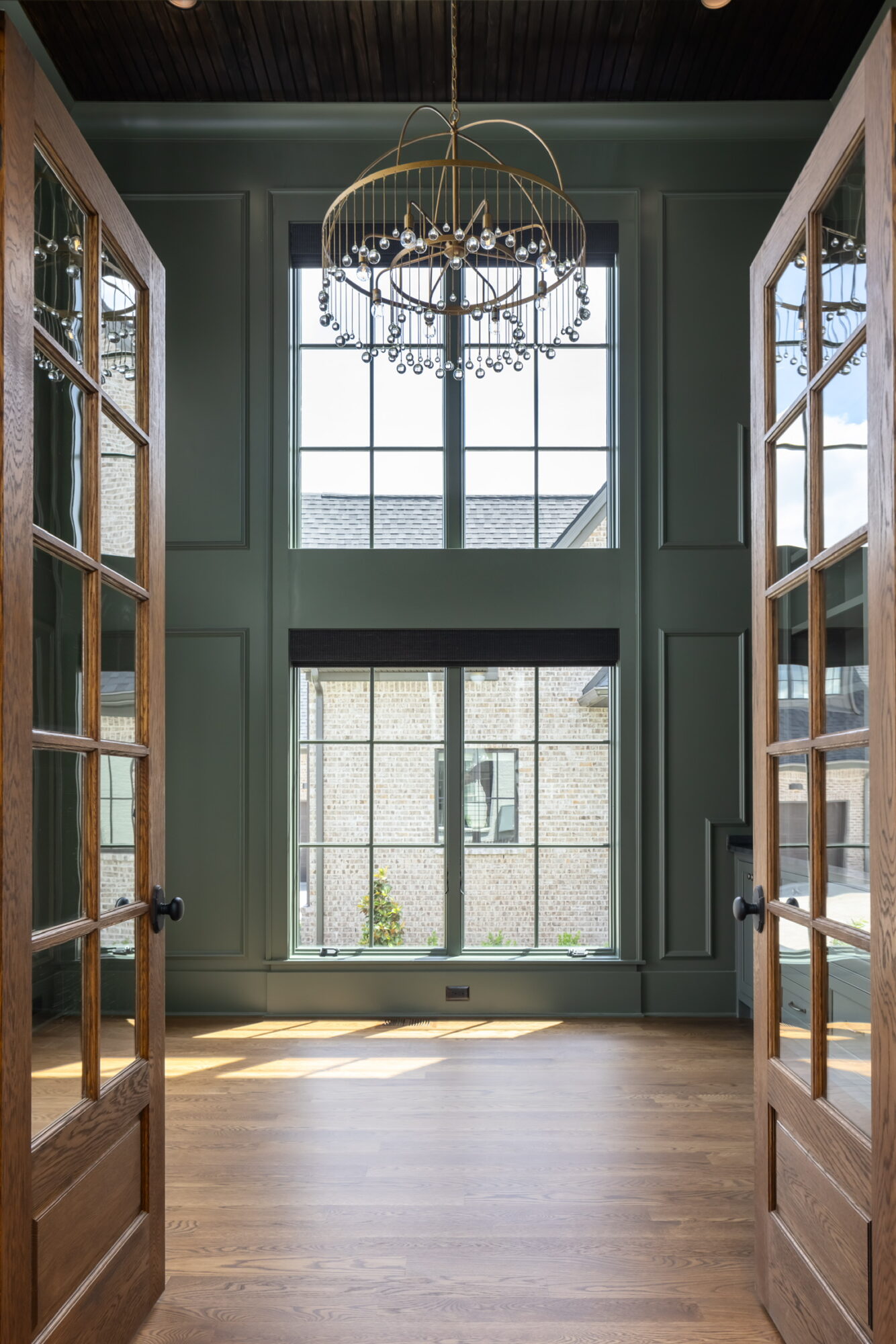 View through open wooden doors into a room with large windows, a chandelier, and wooden flooring.