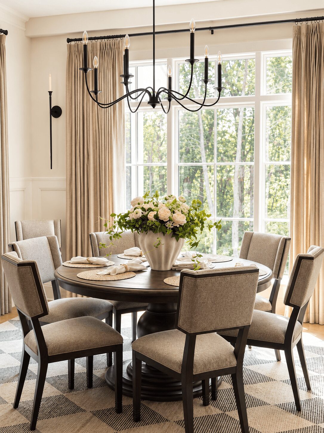 Dining room with a round table, six chairs, a large window with curtains, a chandelier, and a floral centerpiece.