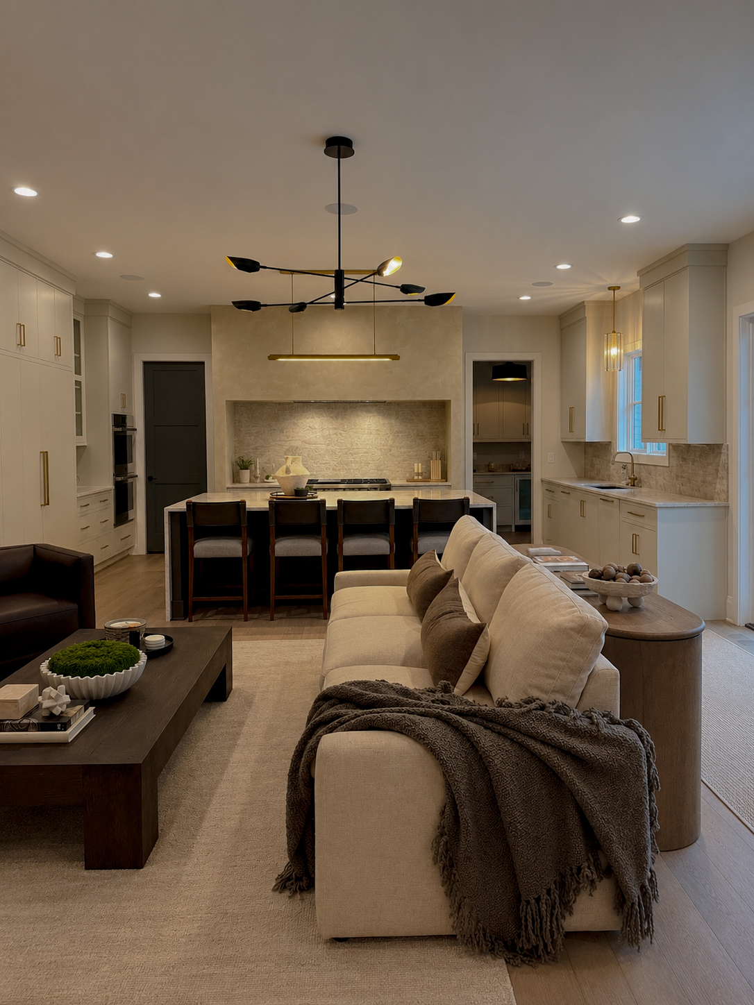 Living room with beige sofa, dark coffee table, and kitchen area with bar stools and a fireplace, illuminated by ceiling lights.