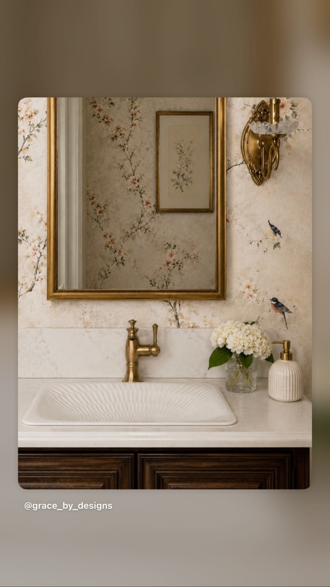 Bathroom sink with mirror, floral wallpaper, and decorative birds on the wall, with a plant and soap dispenser on the counter.