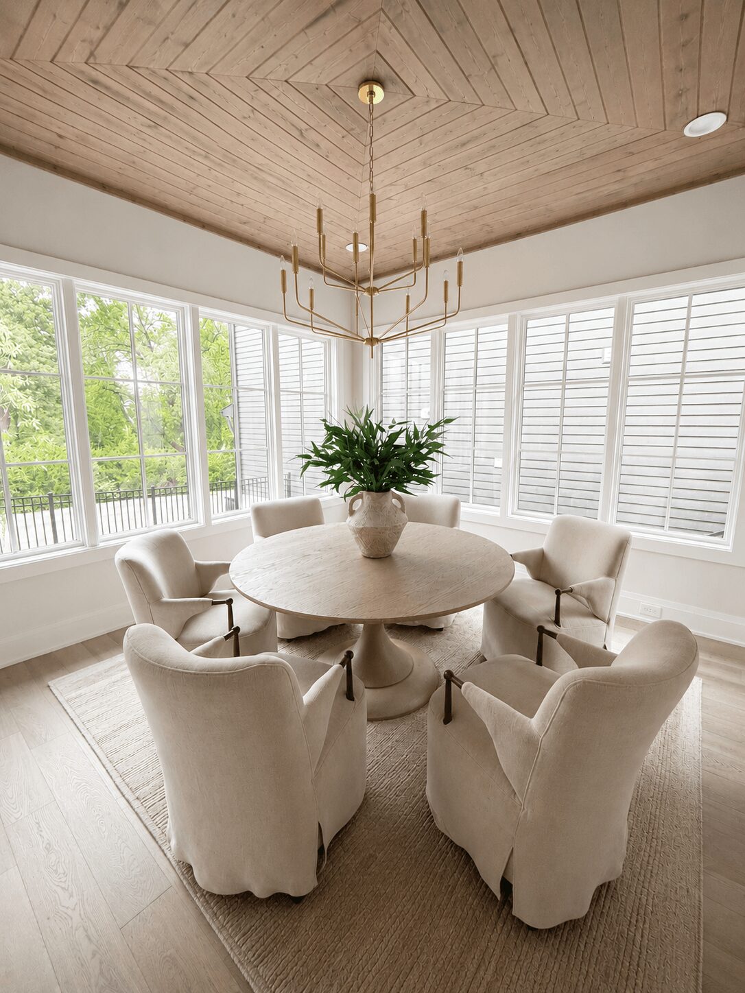 Round dining table with six upholstered chairs and a large potted plant, surrounded by large windows with white blinds, wooden ceiling, and chandelier.