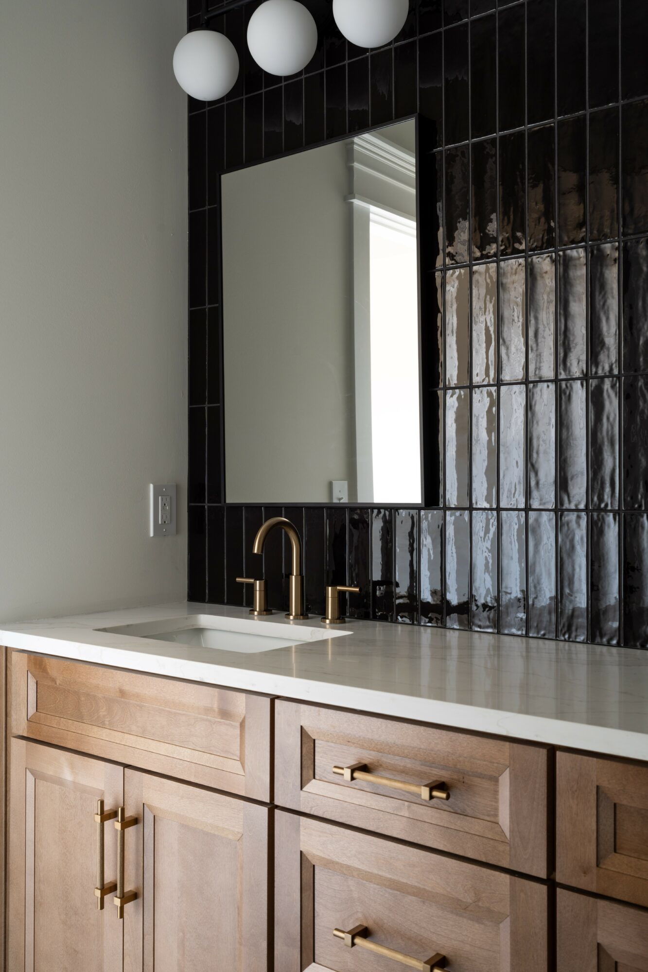 Bathroom vanity with a mirror, black tiled wall, and wooden cabinet with gold handles.