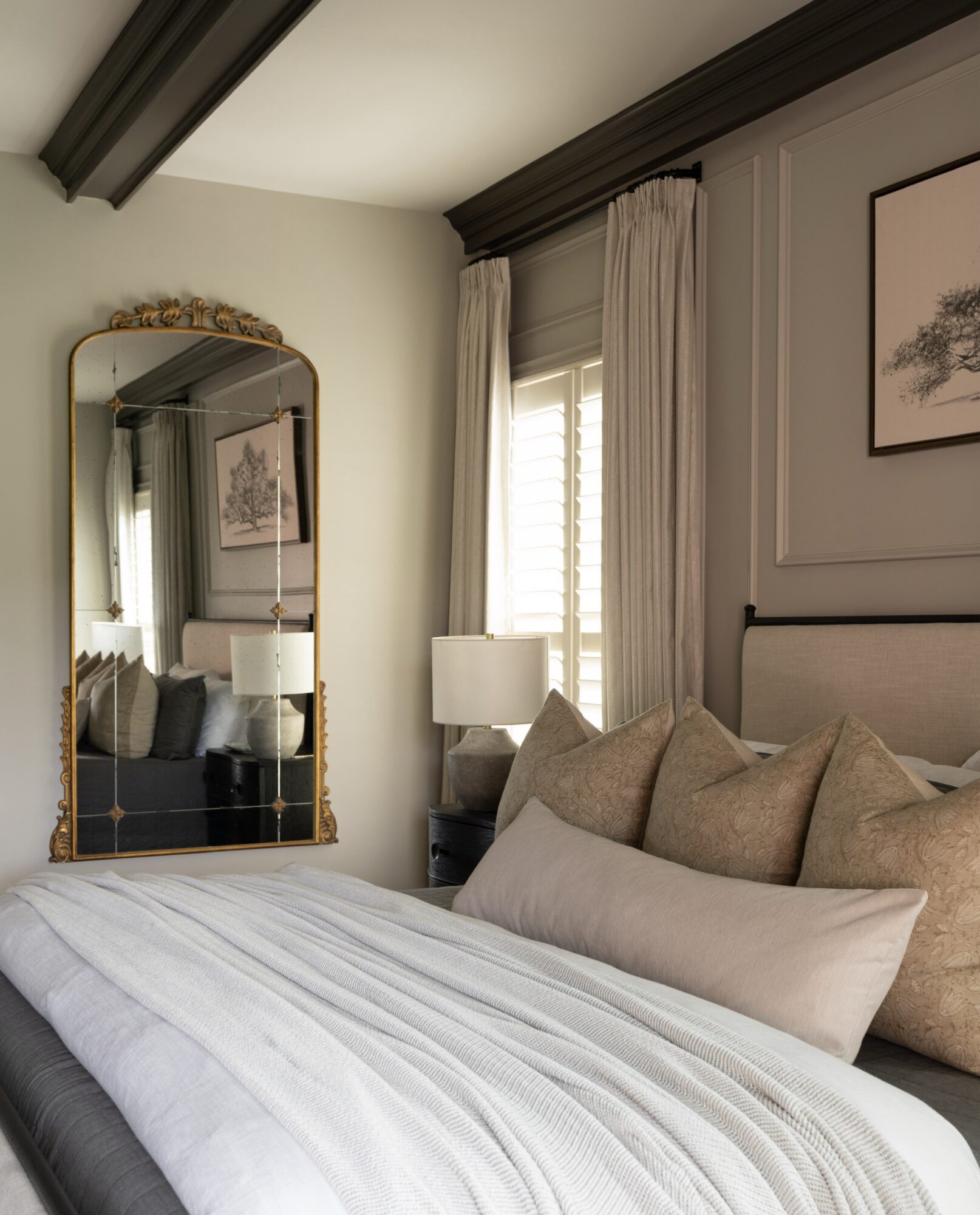Bedroom with bed, pillows, window with curtains, mirror, and framed artwork, in neutral tones.