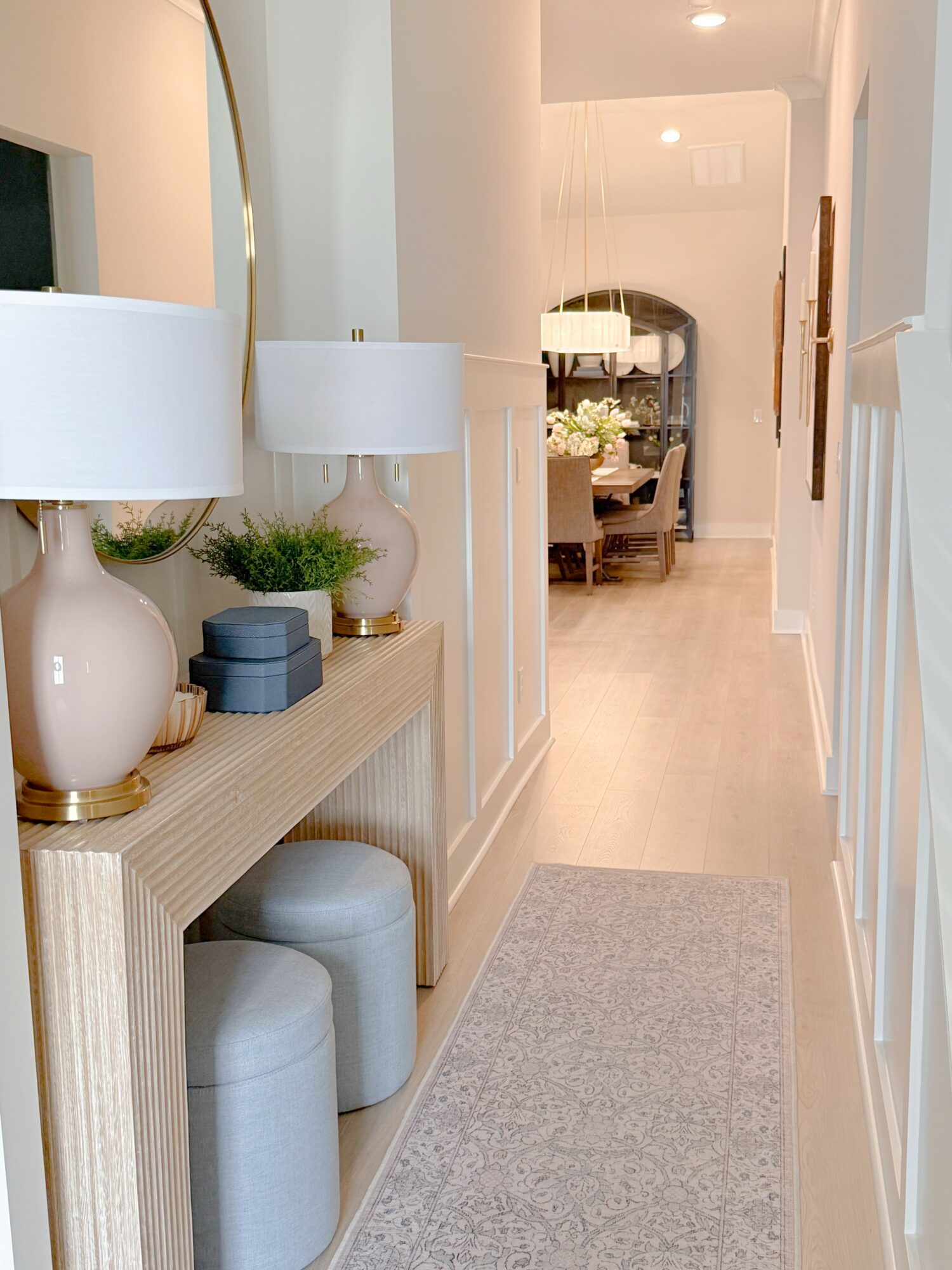 Hallway with console table, lamps, and decorative items leading to dining area, with light-colored walls and flooring.