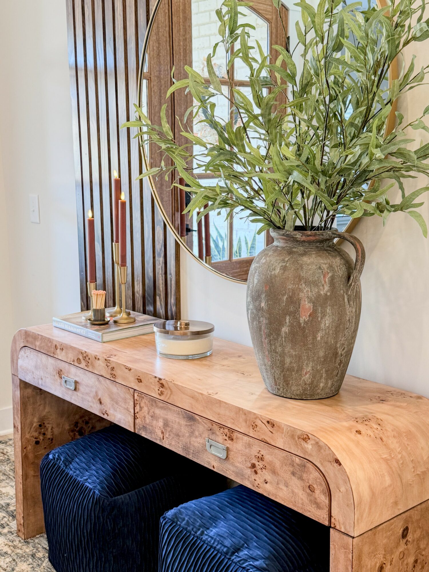 Wooden console table with a large vase of green leafy plant, mirror, candles, and small decorative items, against a wall.