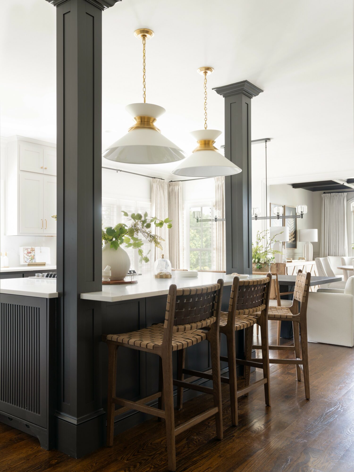 Kitchen with three hanging pendant lights, bar stools at a counter, and a bright, open space with plants and windows.