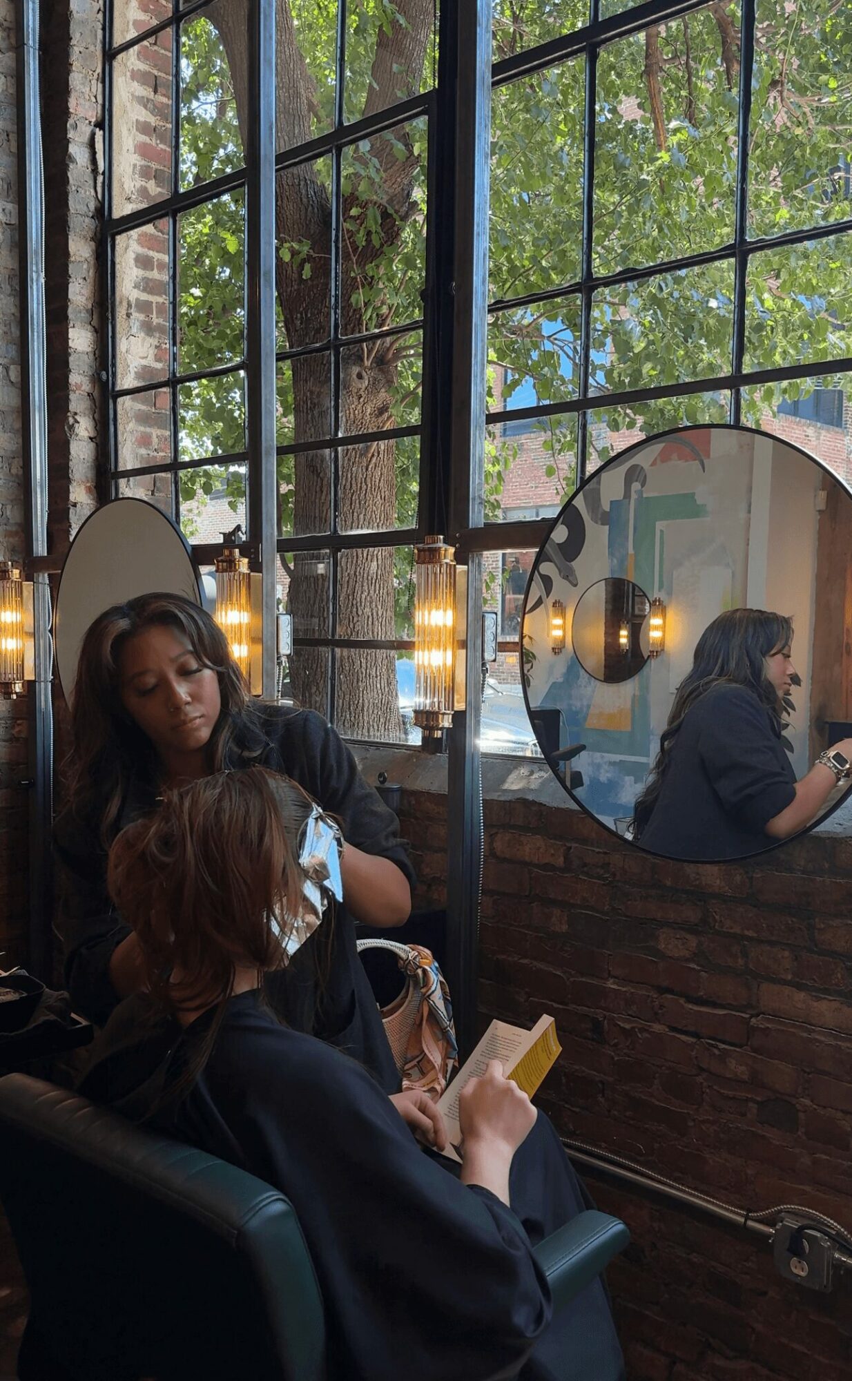 Two women sit in a cafe near a large window with a grid pattern, one looking at her phone, the other in a mirror.