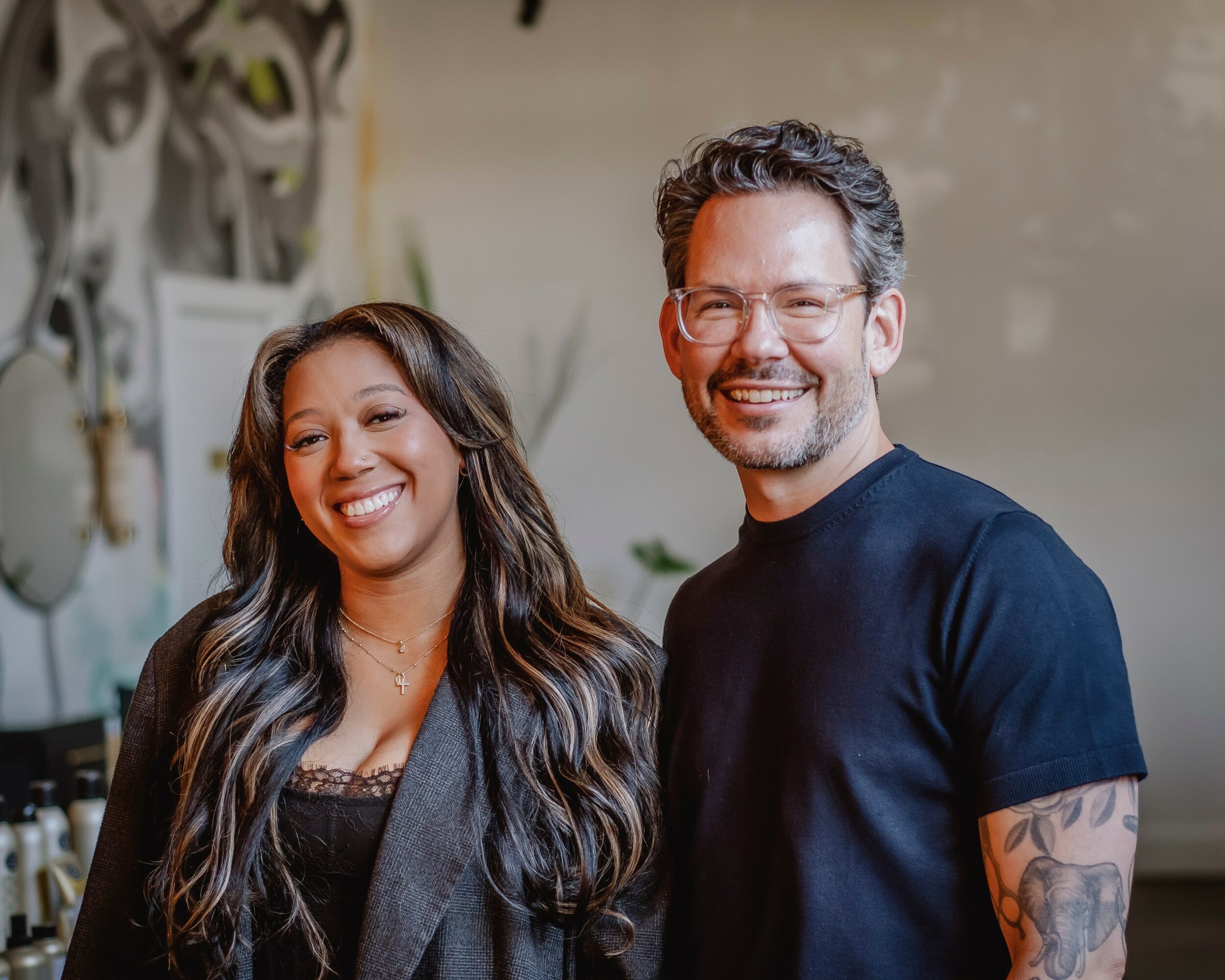 Two smiling people, a woman with long wavy hair and a man with glasses and a tattooed arm, standing indoors.