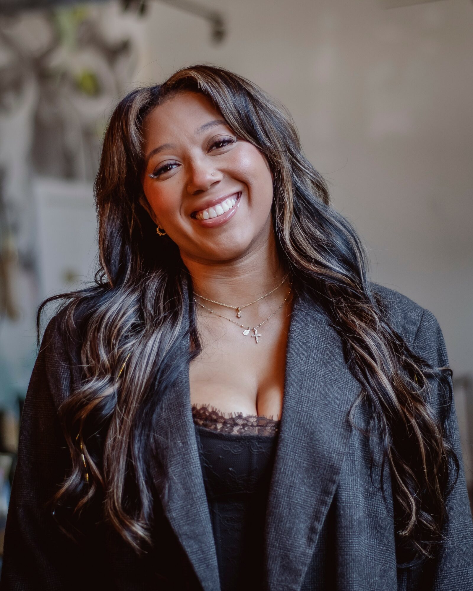 Woman with long wavy hair smiling, wearing a dark blazer and a necklace, standing indoors.