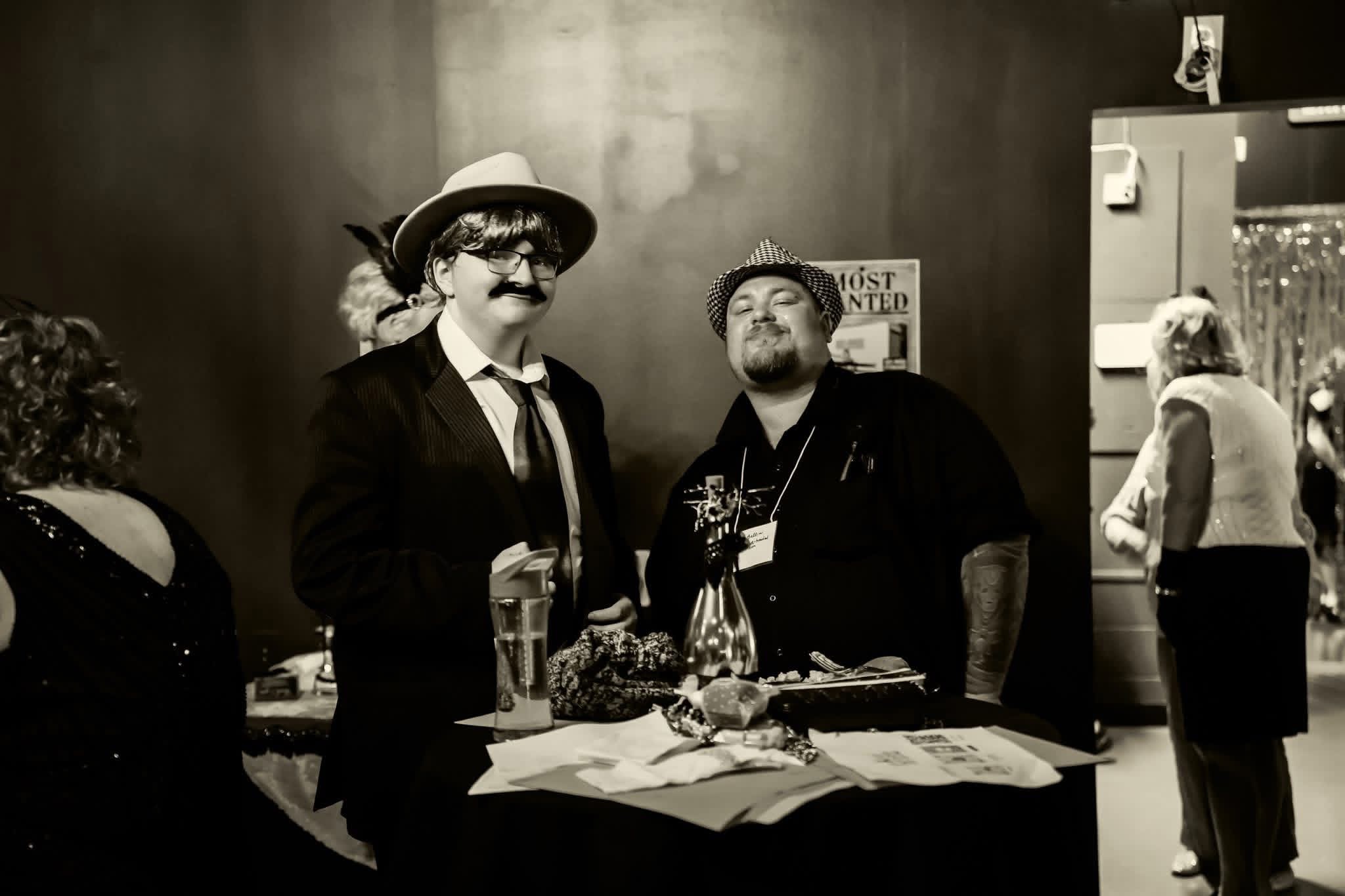 Two men with mustaches and hats stand behind a table, with people in the background, in a black-and-white setting.