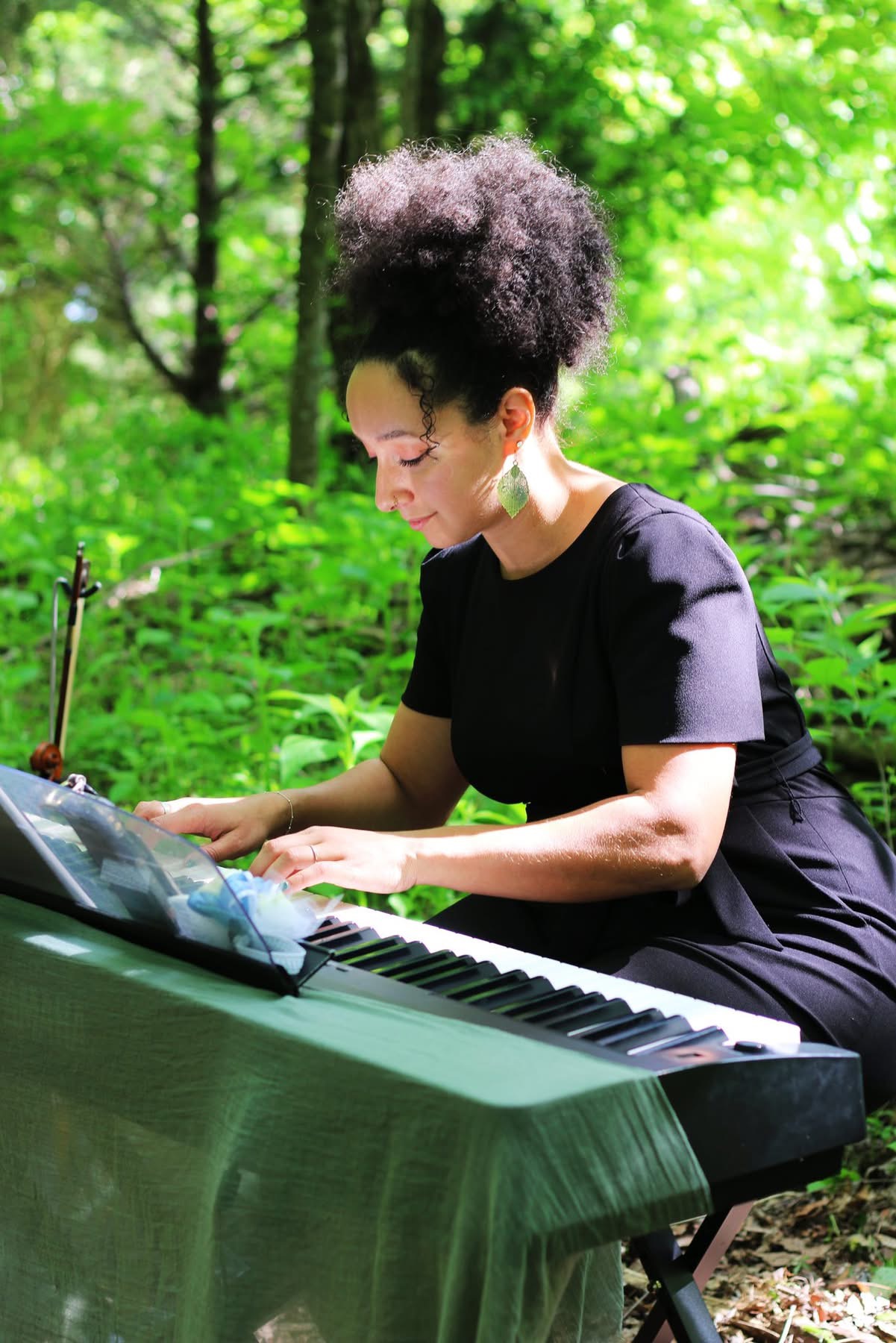 Woman with curly hair playing a keyboard outdoors in a green forest, smiling and focused.