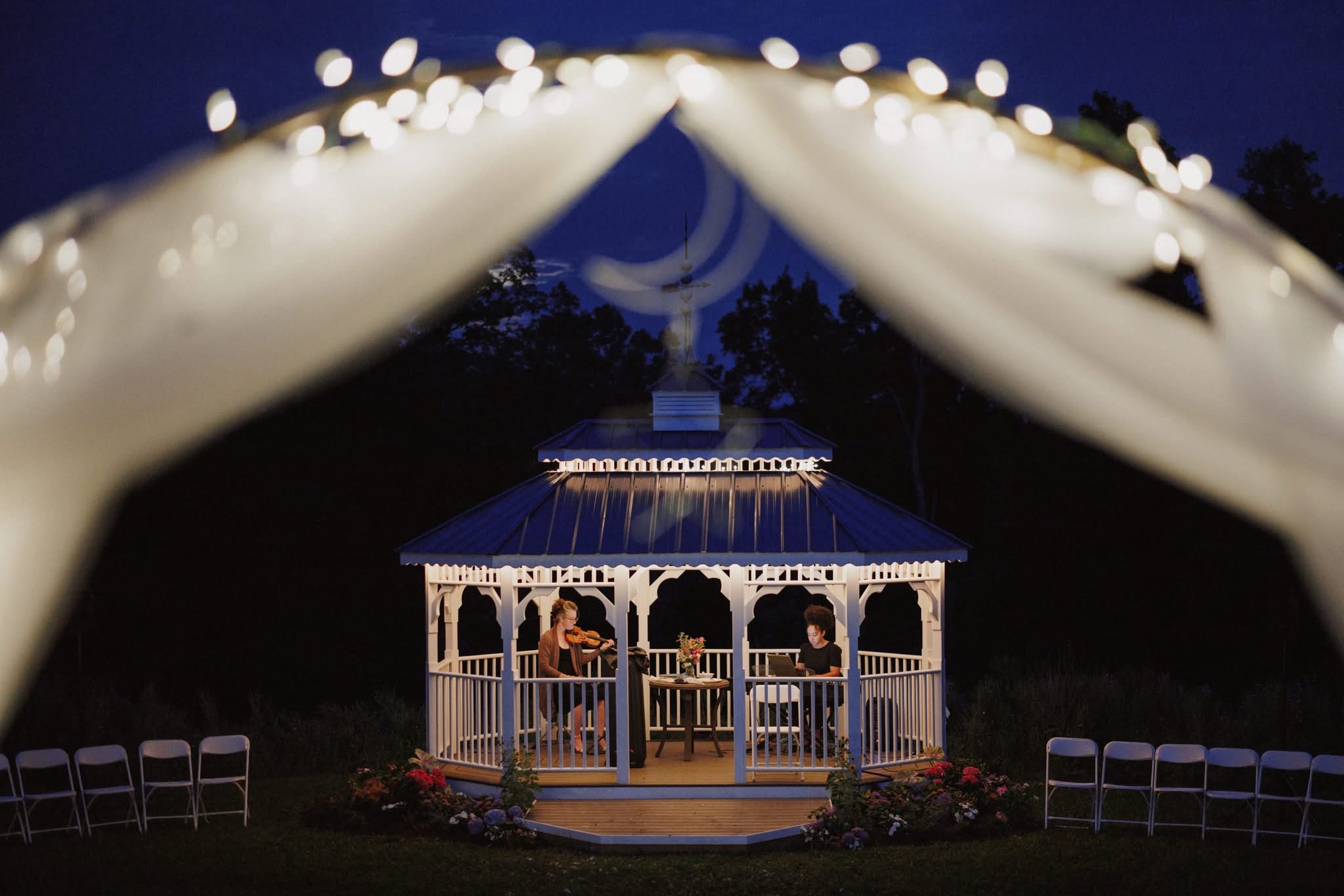 Outdoor gazebo with people inside, decorated with string lights, under a night sky with a crescent moon, surrounded by chairs.