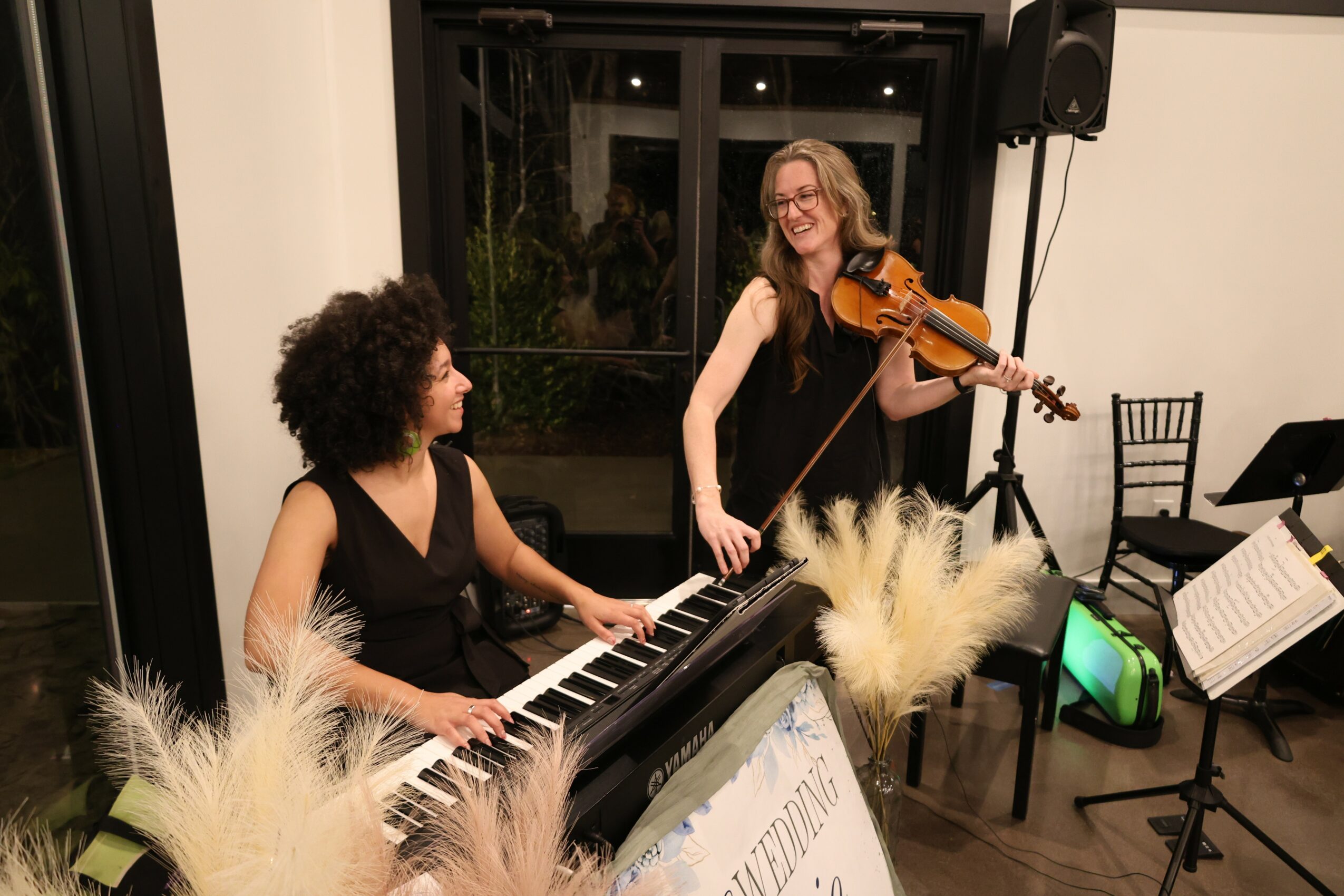 Two women playing musical instruments indoors, one on keyboard and the other on violin, smiling and enjoying music, with music stands nearby.