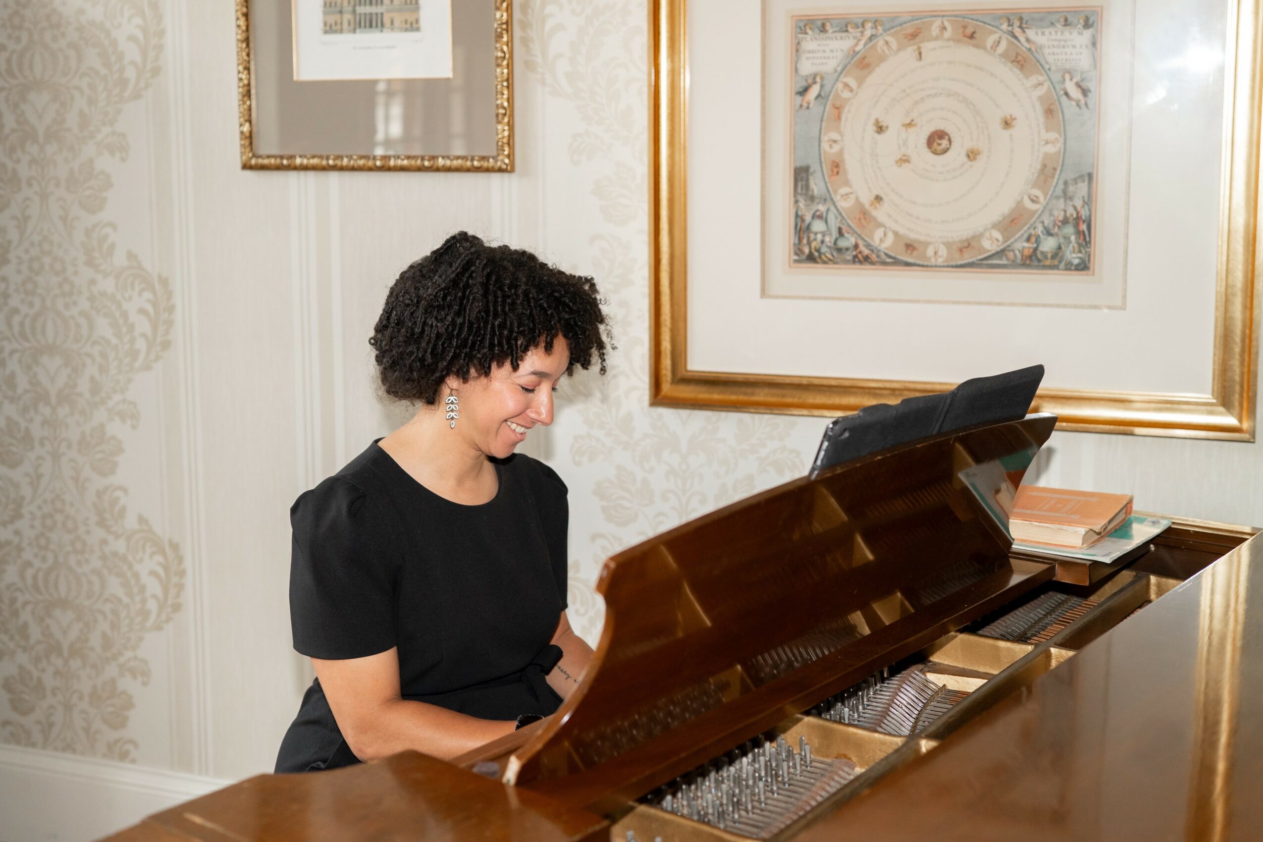Woman with curly hair playing a grand piano in a room with framed artwork and ornate wallpaper.