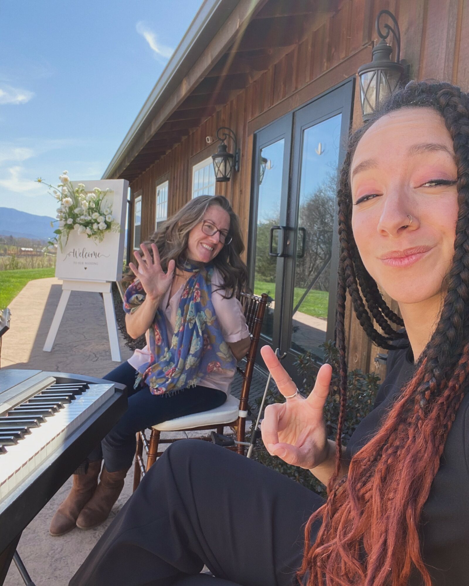 Two women smiling outdoors, one making a peace sign, with a piano and wooden building in background.