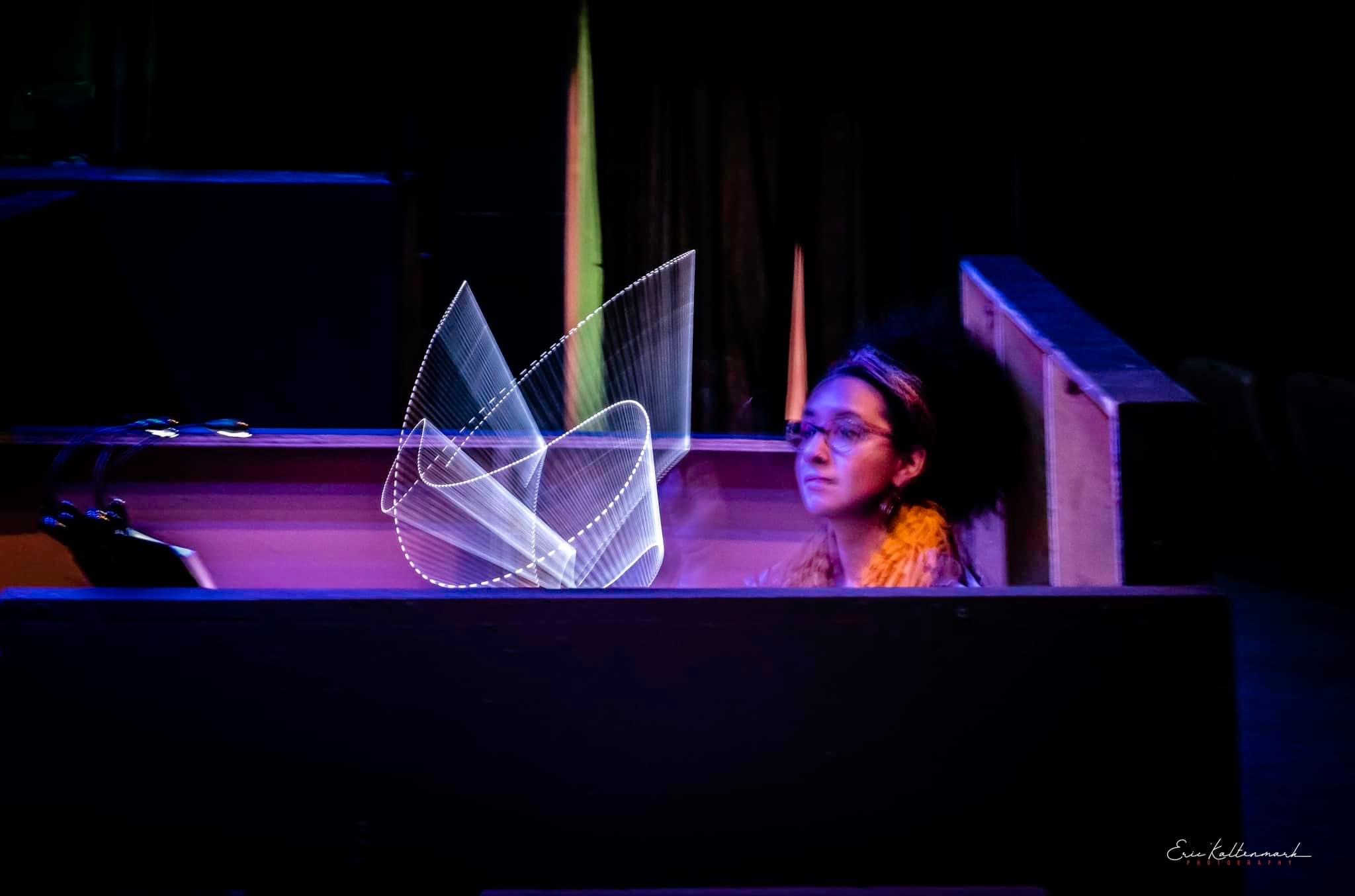 Young girl with glasses sitting at a piano with a transparent sculpture, dark background, stage setting.