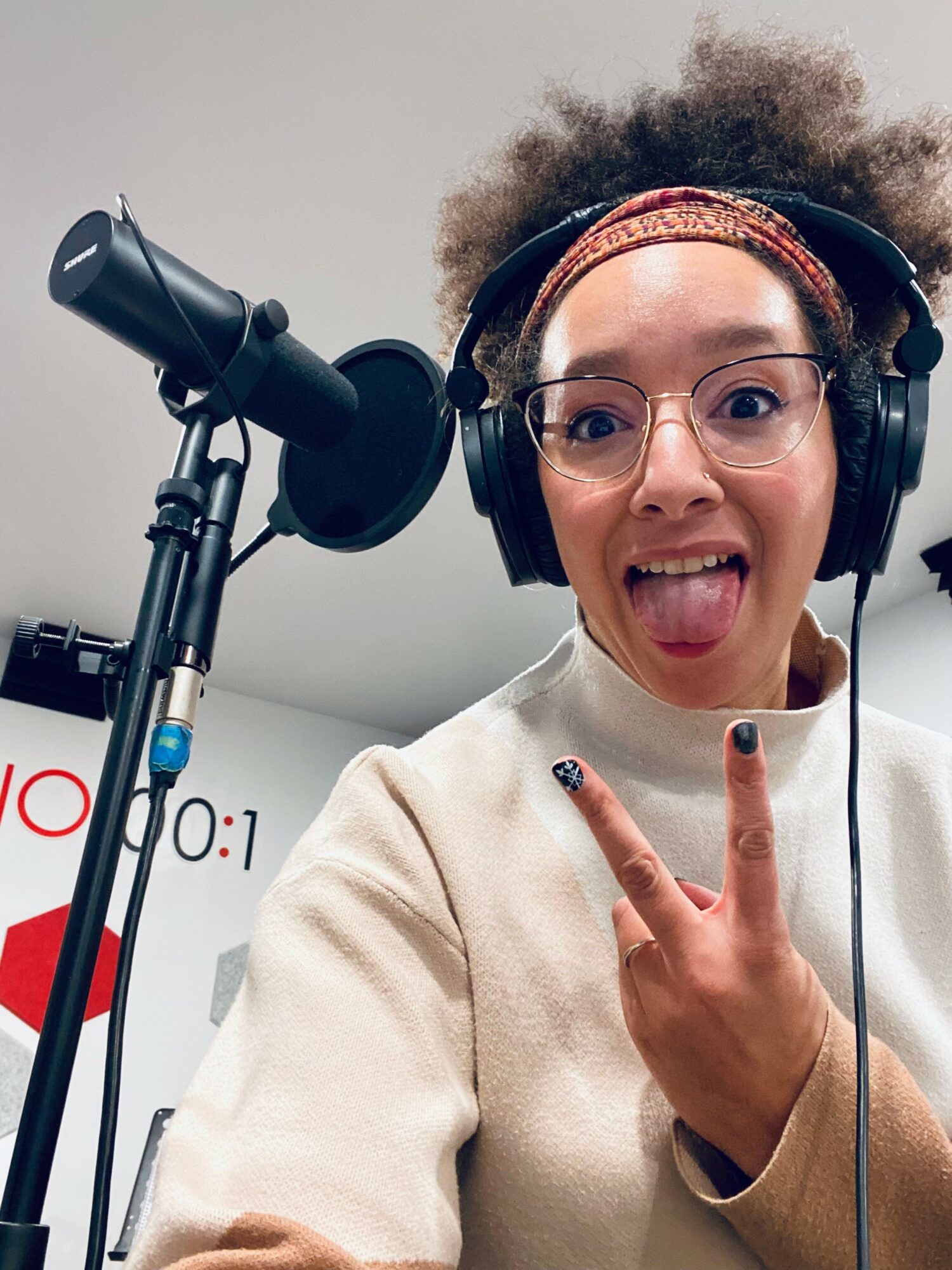 Young woman with glasses and headphones making a peace sign and sticking out her tongue in front of a microphone.