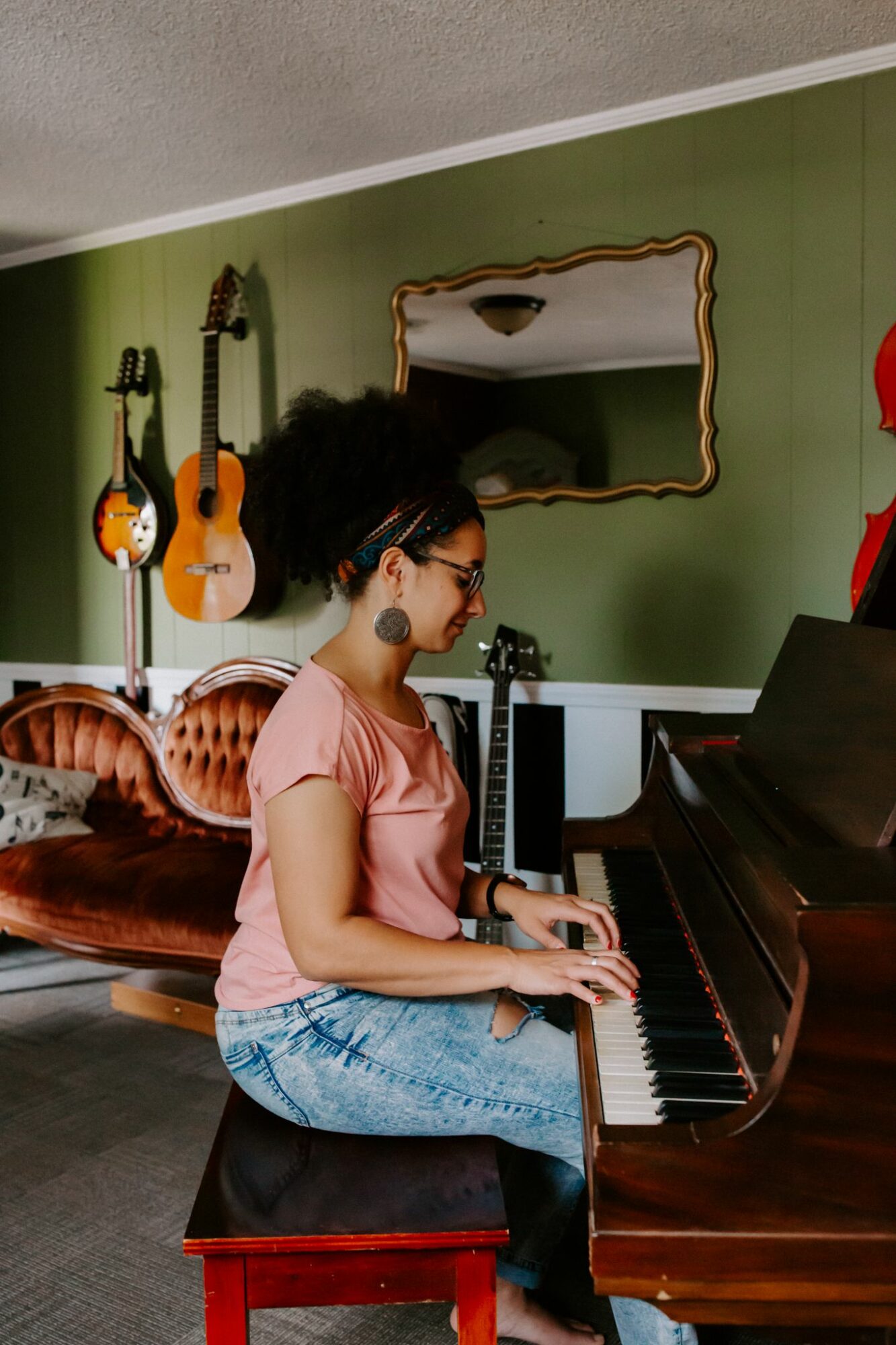 Young woman playing piano in a room with green walls, guitars hanging, and a mirror, sitting on a stool.