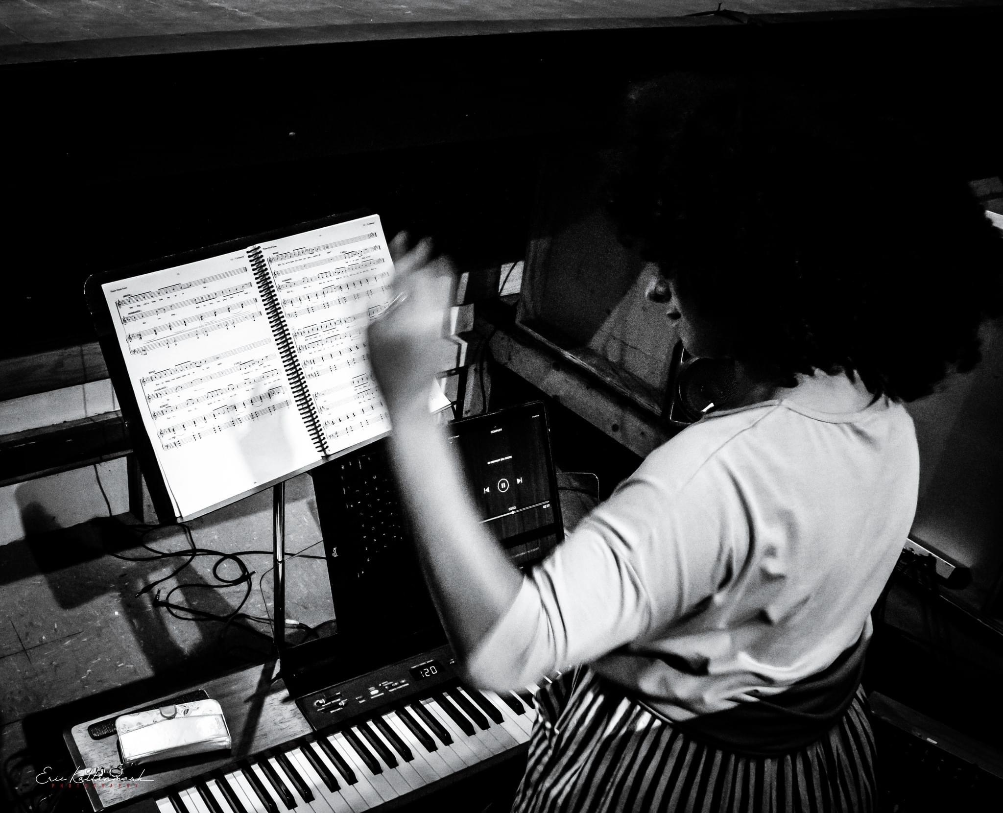 Person with curly hair conducting music with sheet music and keyboard, in a dark room.