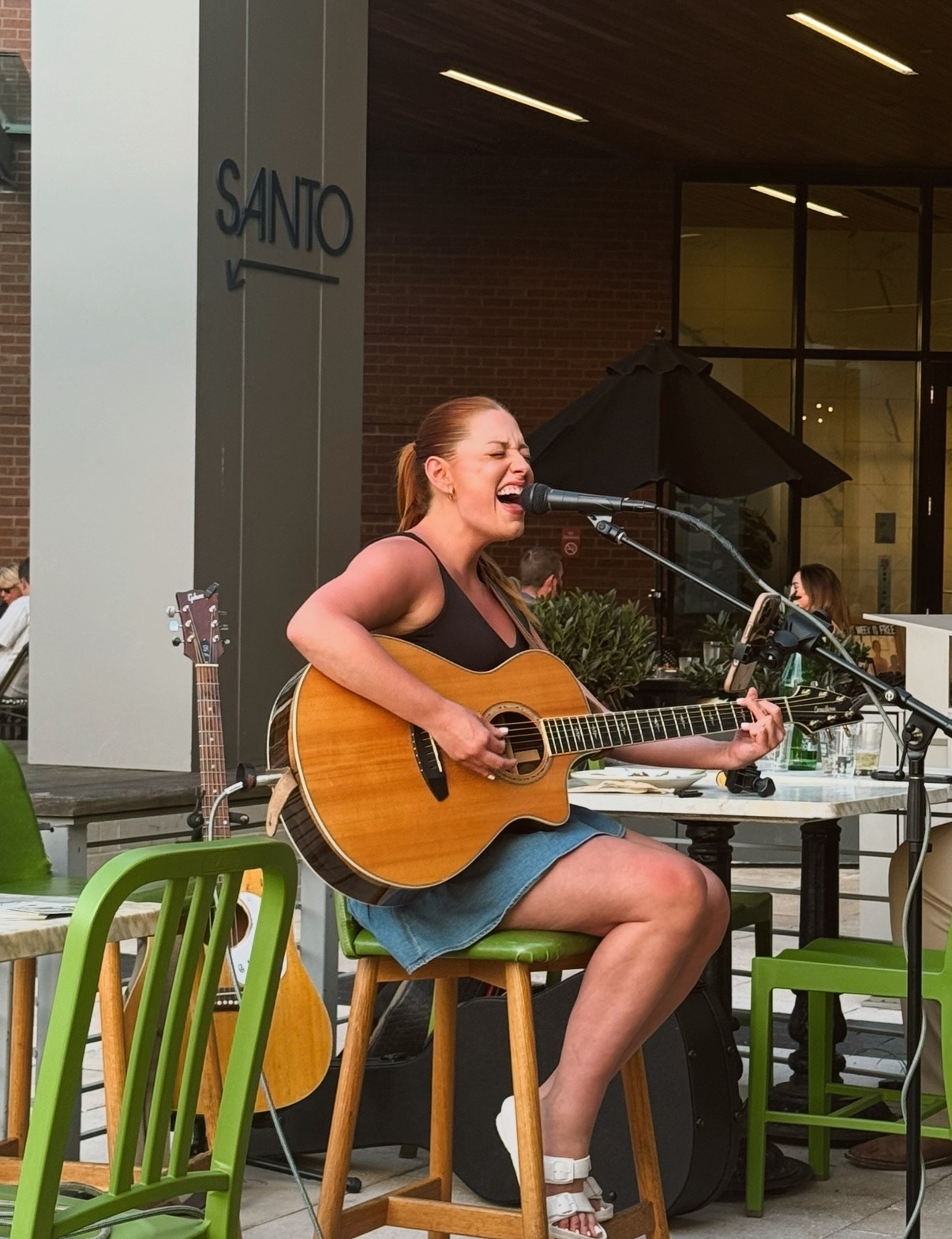 Woman playing guitar and singing outdoors, sitting on a green chair, with a building and outdoor seating in background.