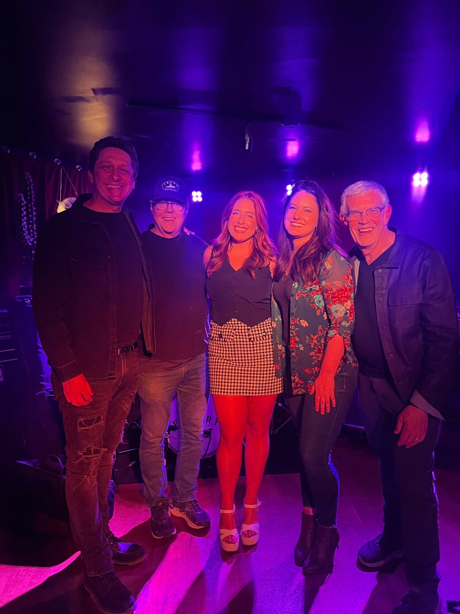 Group of six people standing together in a dark setting with purple lighting, some smiling and facing the camera.