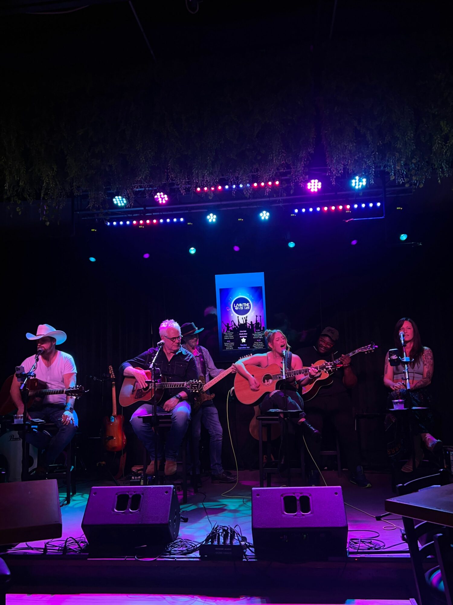 Musicians perform on stage with colorful lights and a screen in the background, playing instruments and singing.