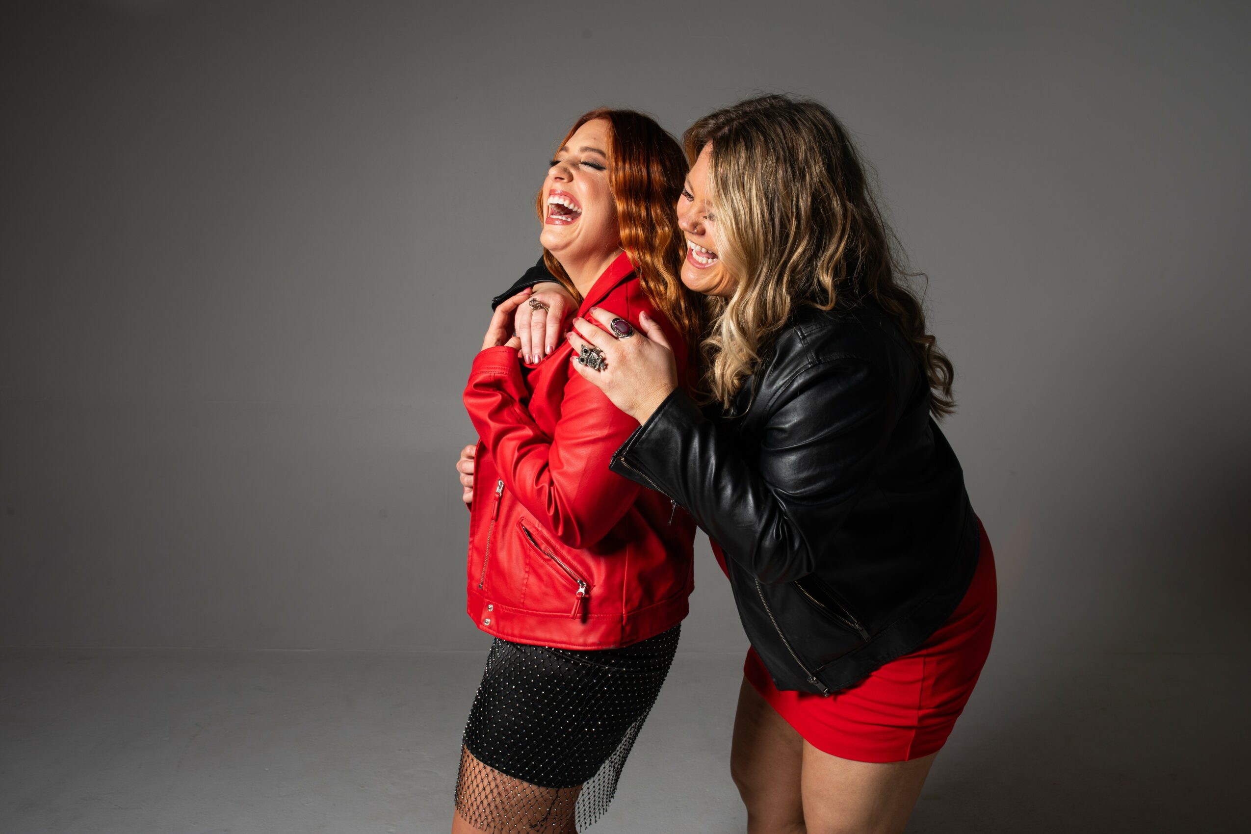 Two women laughing and hugging, one in a red jacket and black skirt, the other in a black jacket and red skirt, against a gray background.