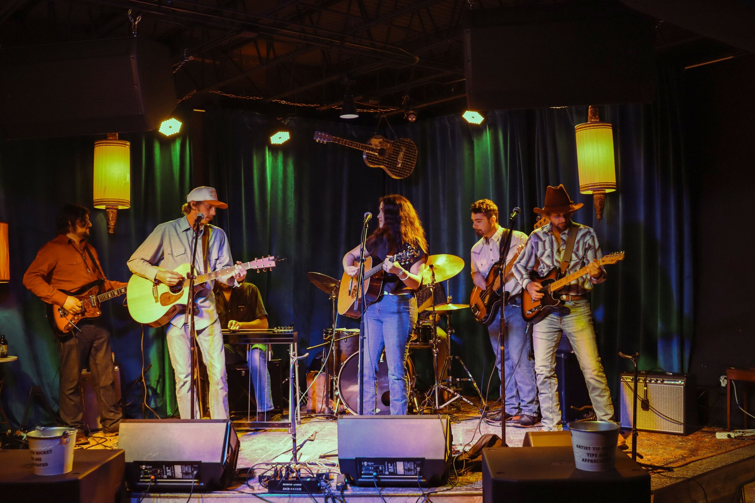 Five musicians perform on stage with guitars and drums, colorful lighting, and a dark curtain backdrop.