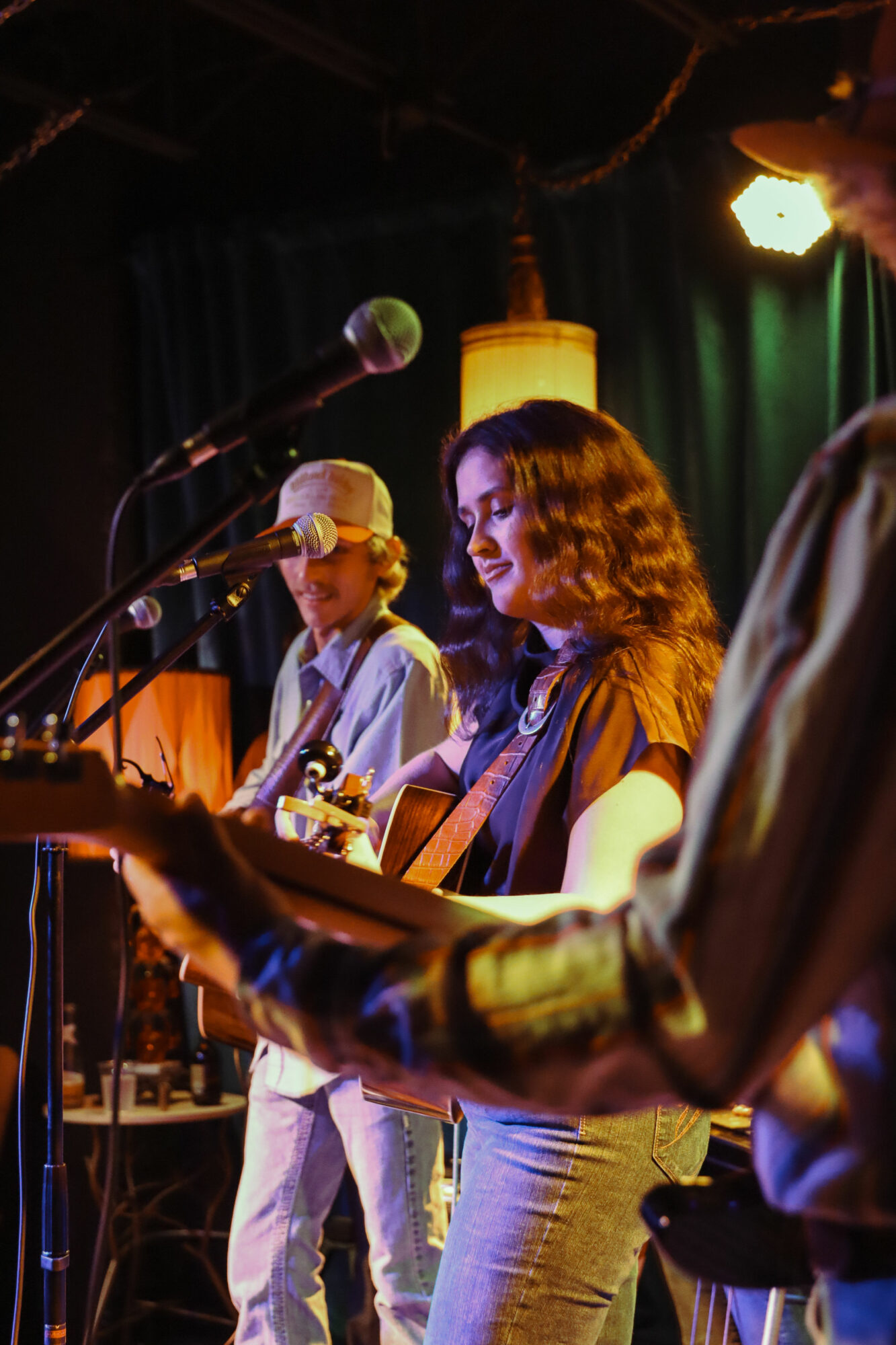 Three musicians perform on stage with microphones, guitar, and a person in the foreground playing an instrument, dimly lit background.