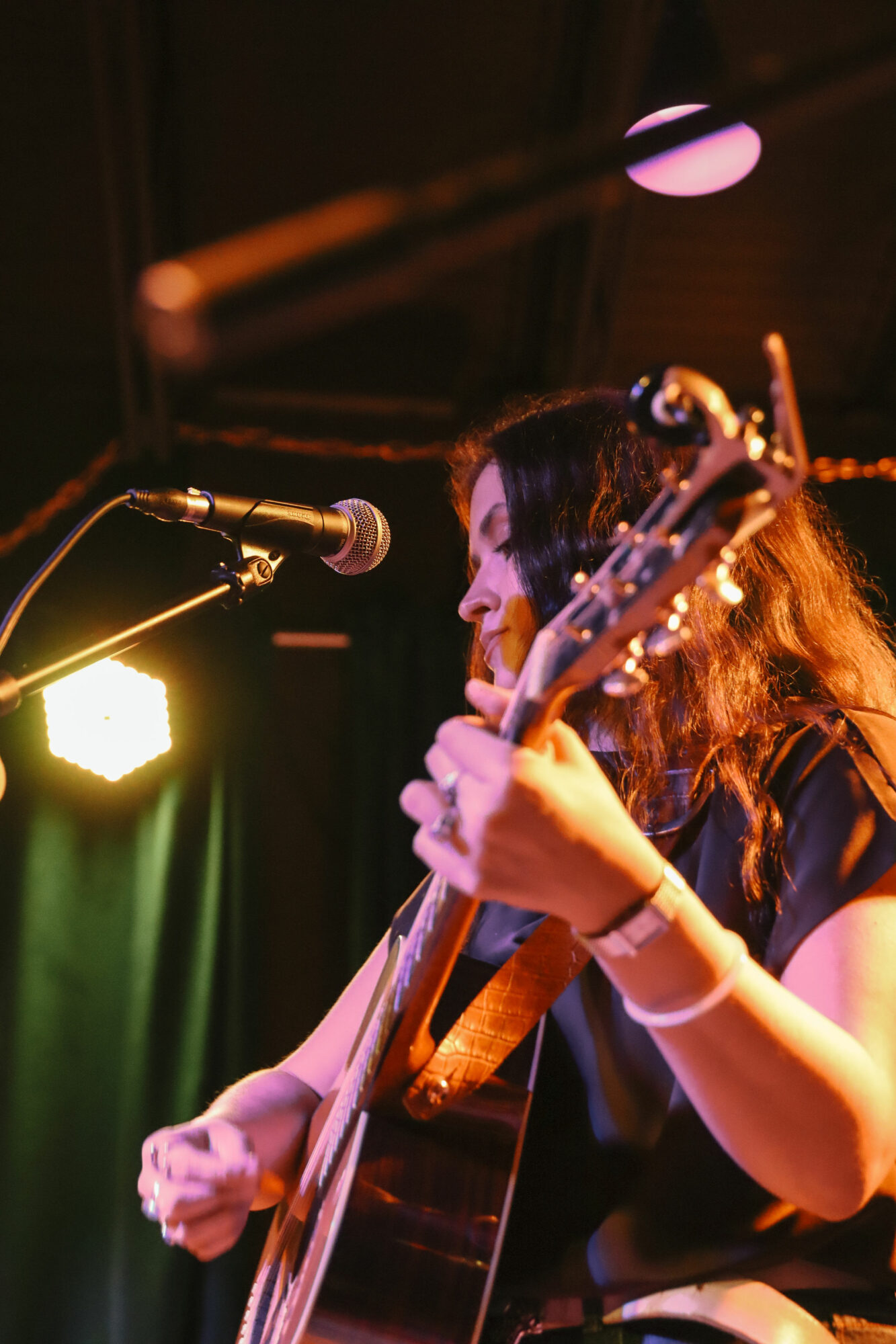 Female musician playing guitar and singing into a microphone on stage with colorful lighting.