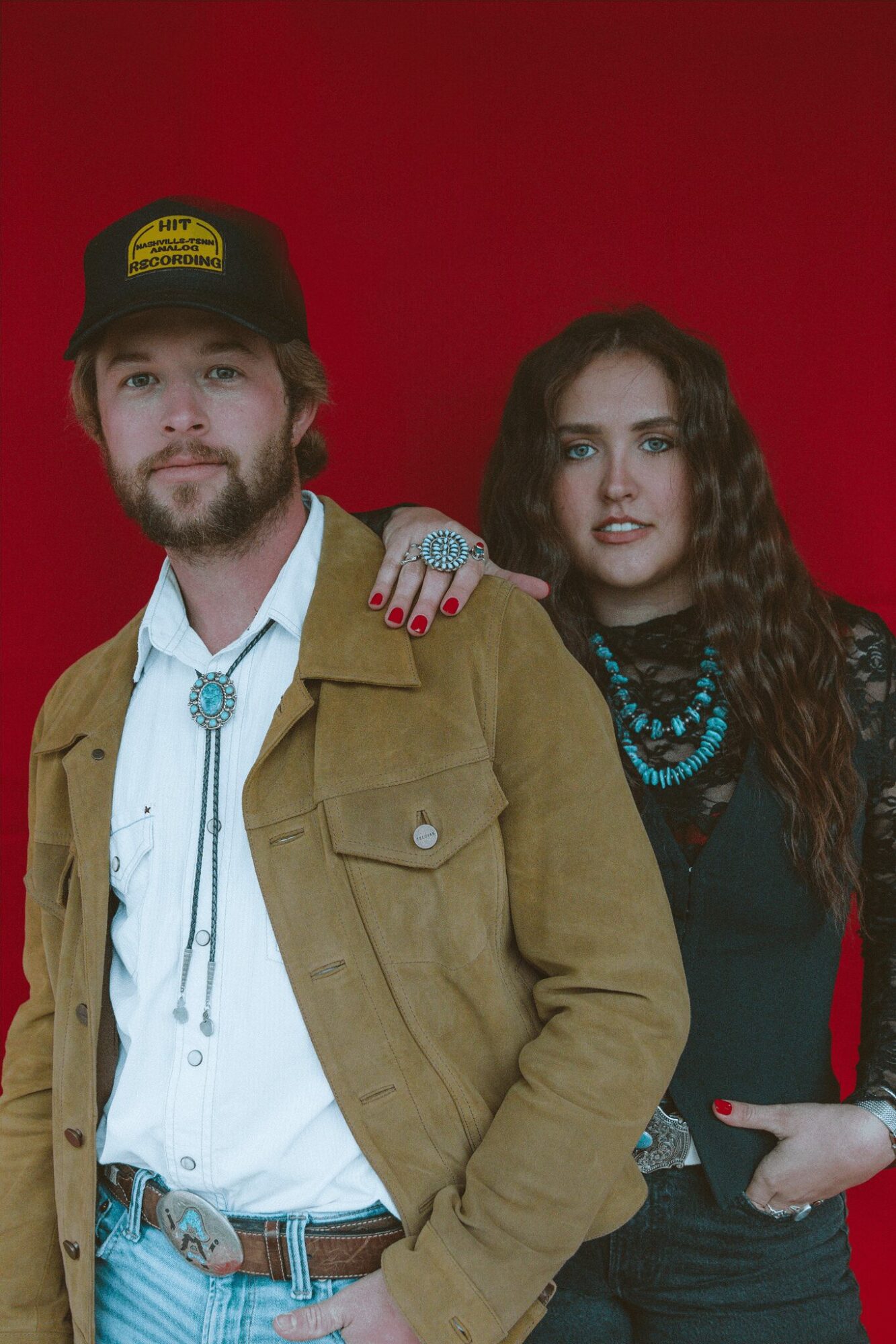 A man and woman stand close together against a red background, with the woman's hand on the man's shoulder.