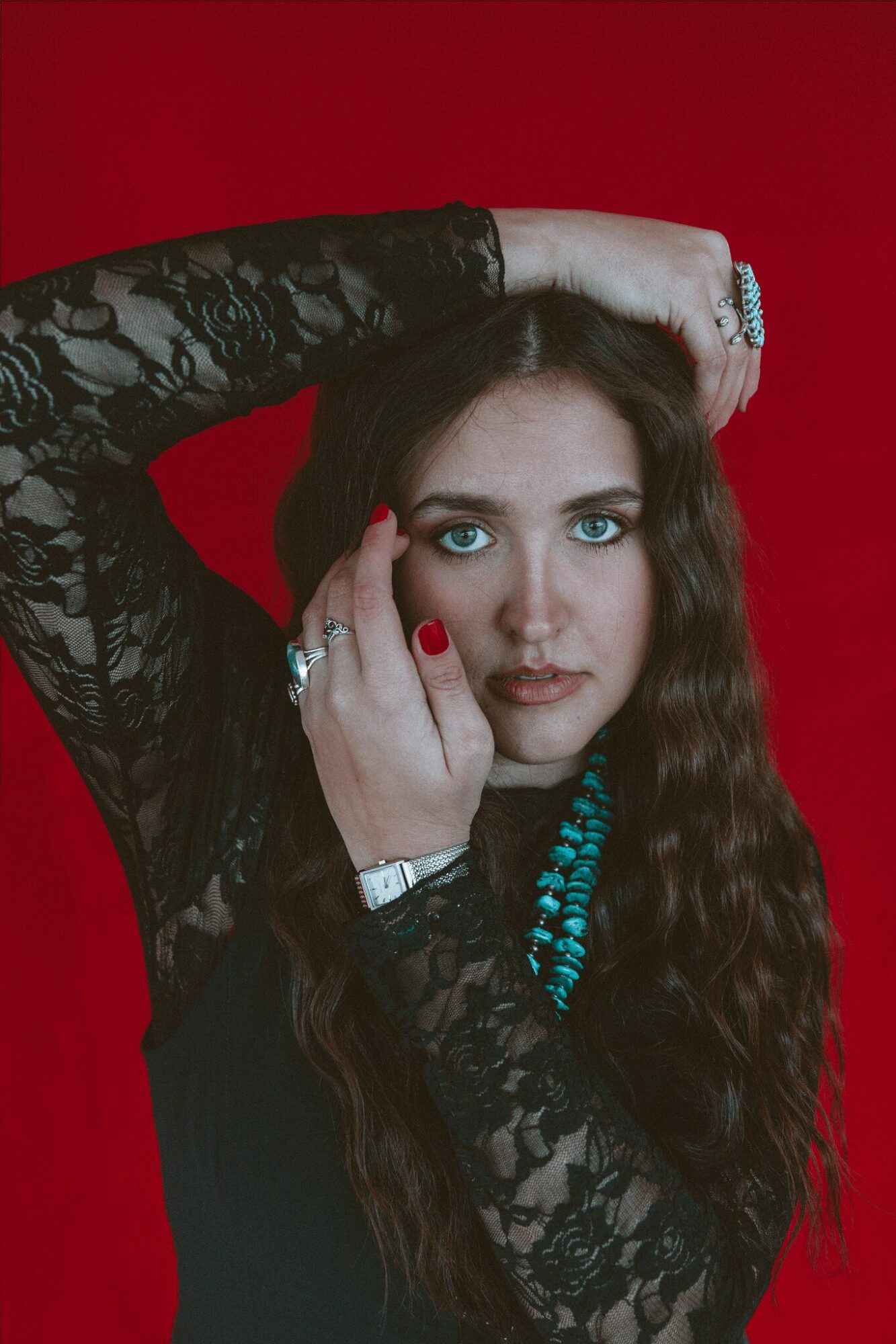 Woman with long wavy hair, wearing black lace top, turquoise necklace, and red lipstick, posing against red background.